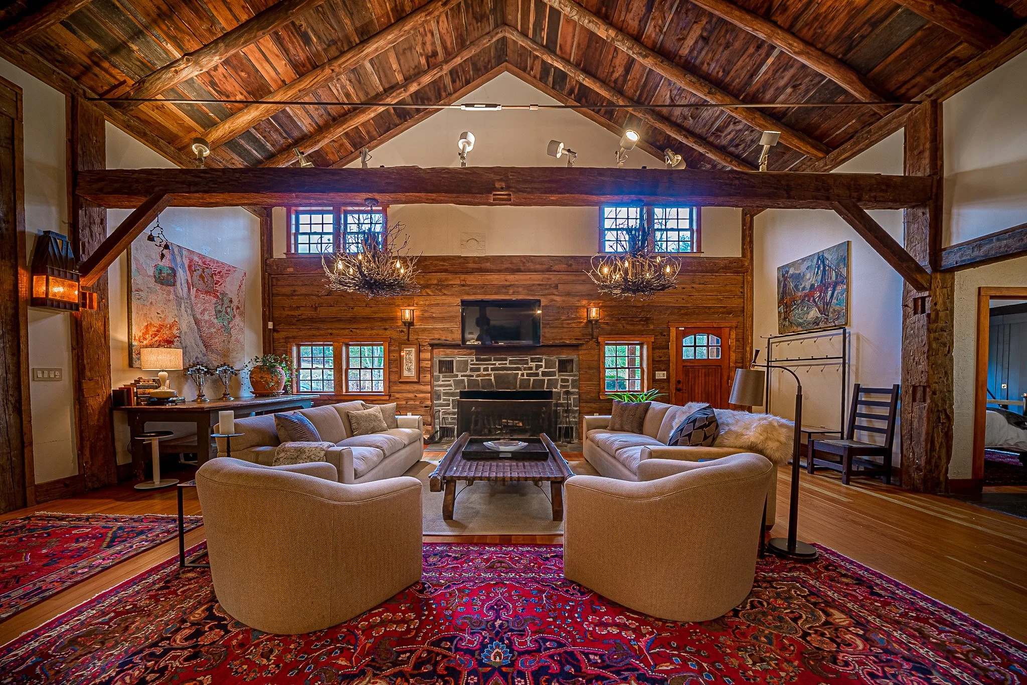 A rustic living room with wooden walls and a vaulted wood ceiling, furnished with beige and cream sofas, a wooden coffee table, and a patterned red and blue area rug. There is a fireplace with a stone surround, a flat-screen TV mounted above it, and