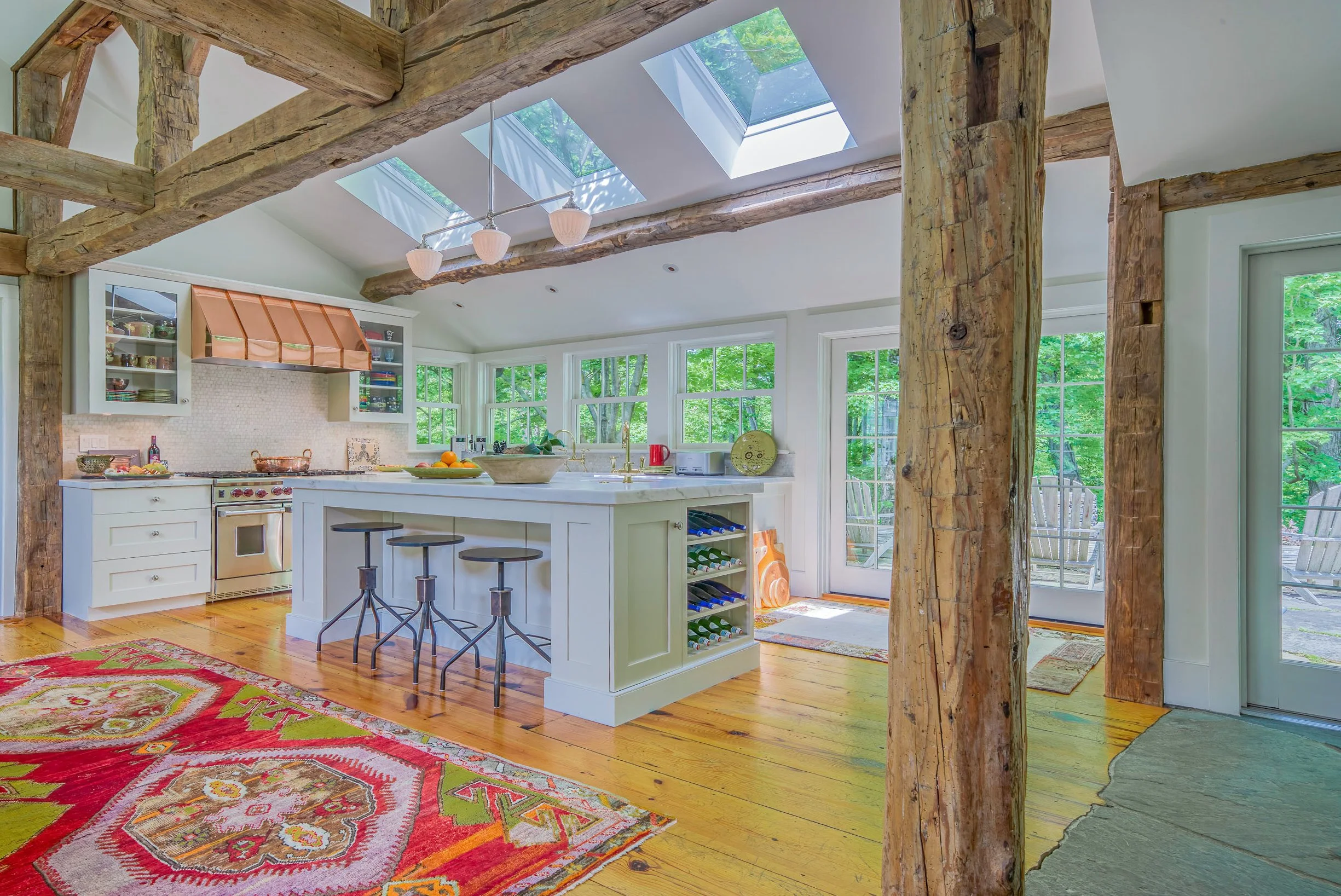 Bright kitchen with white cabinetry, wood beams, skylights, and large windows overlooking a green outdoor area.