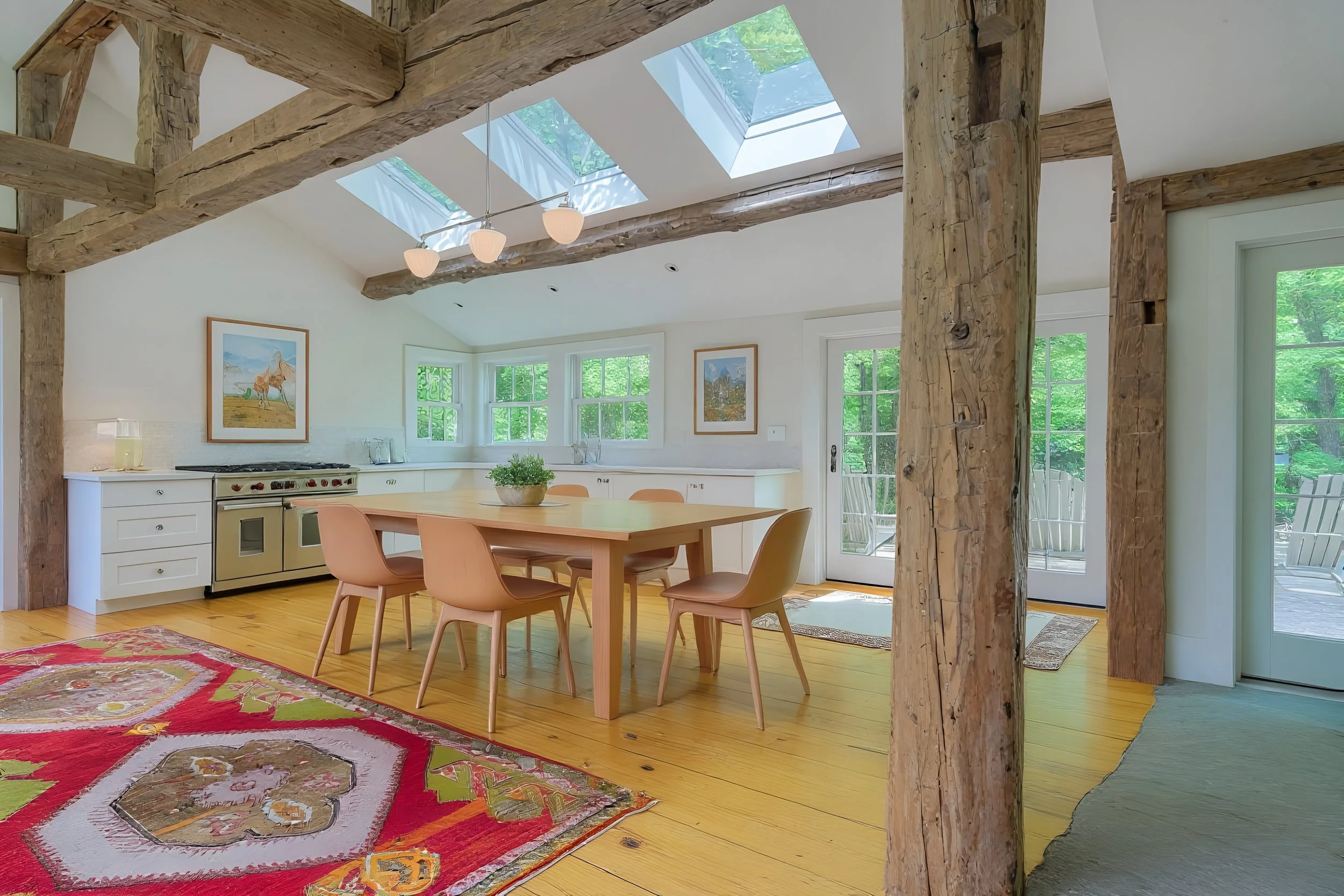 Bright kitchen and dining area with wooden beams, skylights, white cabinets, a wooden dining table with six chairs, and a colorful area rug. Large windows and a glass door lead to an outdoor patio with green trees outside.