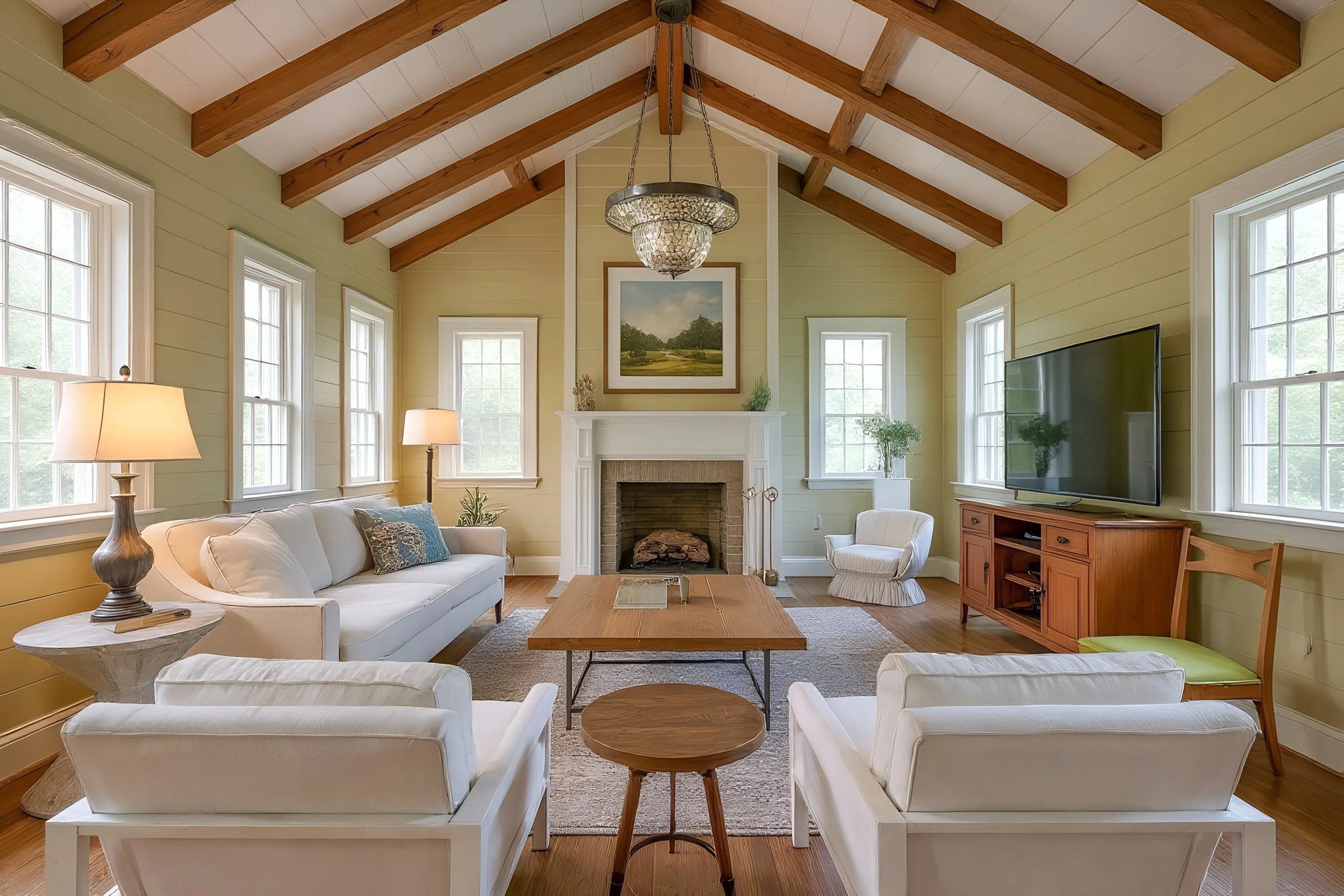 Living room with vaulted ceiling, exposed wooden beams, large windows, white sofa, chairs, wooden coffee table, fireplace, TV, and various lamps and plants.