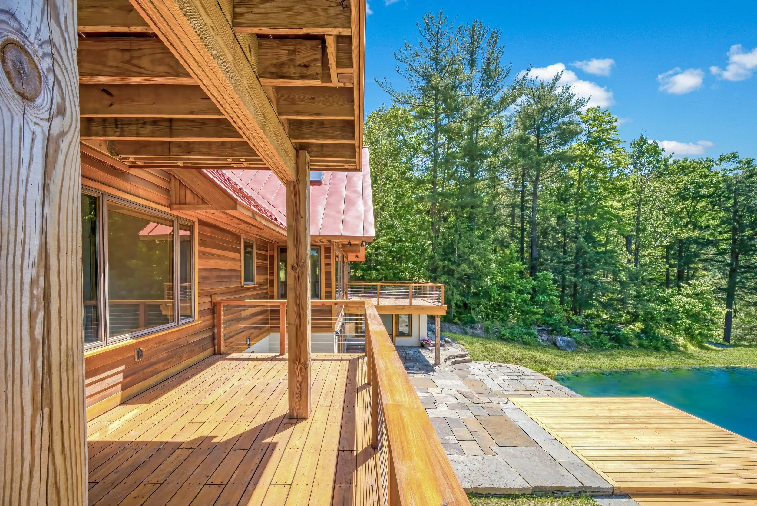 Wooden deck and balcony attached to a house with a red metal roof, overlooking a pond and a lush green forest under a blue sky.