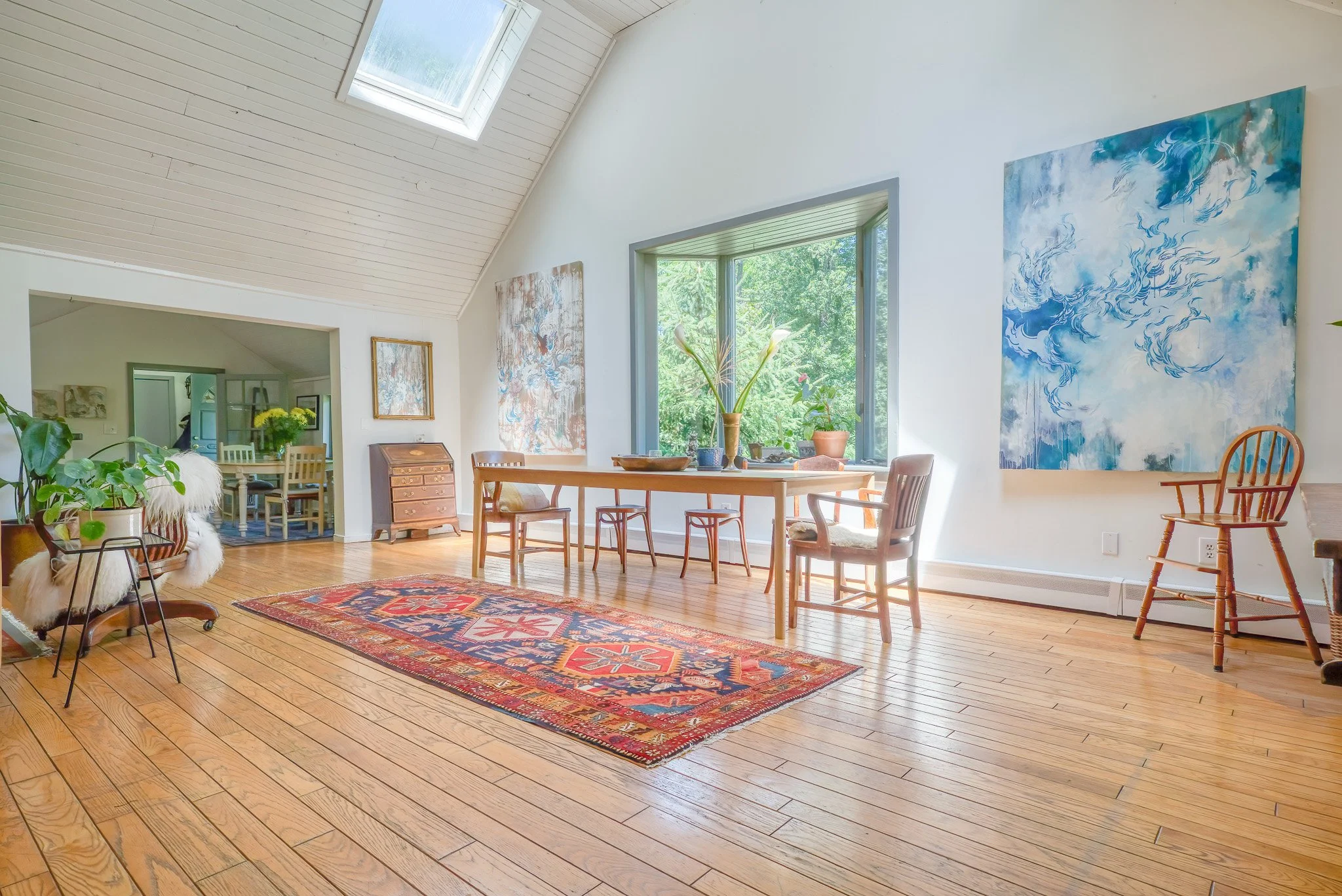 Bright dining room with wooden flooring, large window, colorful abstract paintings, a wooden table with chairs, and a rug, with a view of a lush green outdoor area.