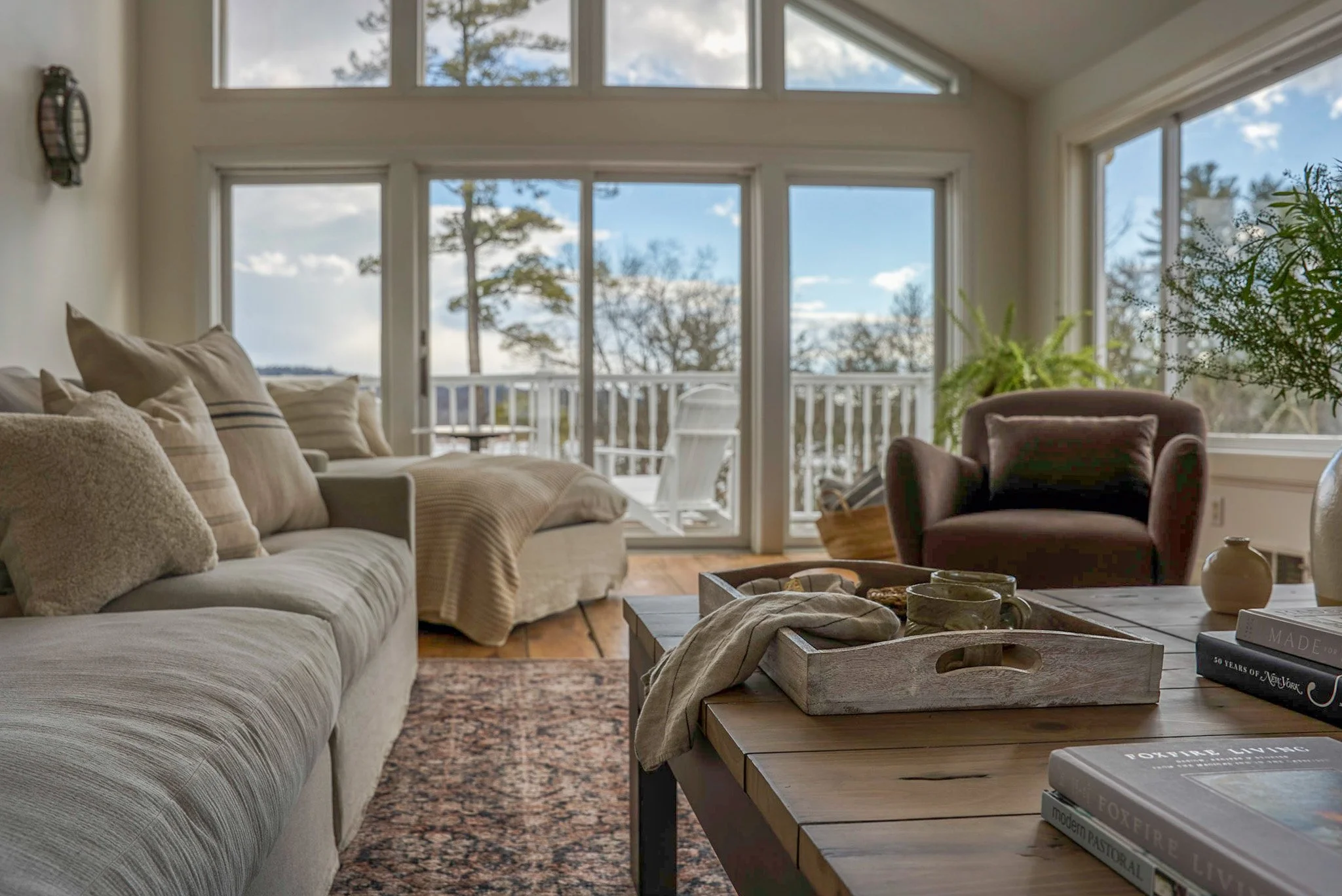 Living room with beige sofa, armchair, coffee table with books, and large windows showing trees and a balcony outside.