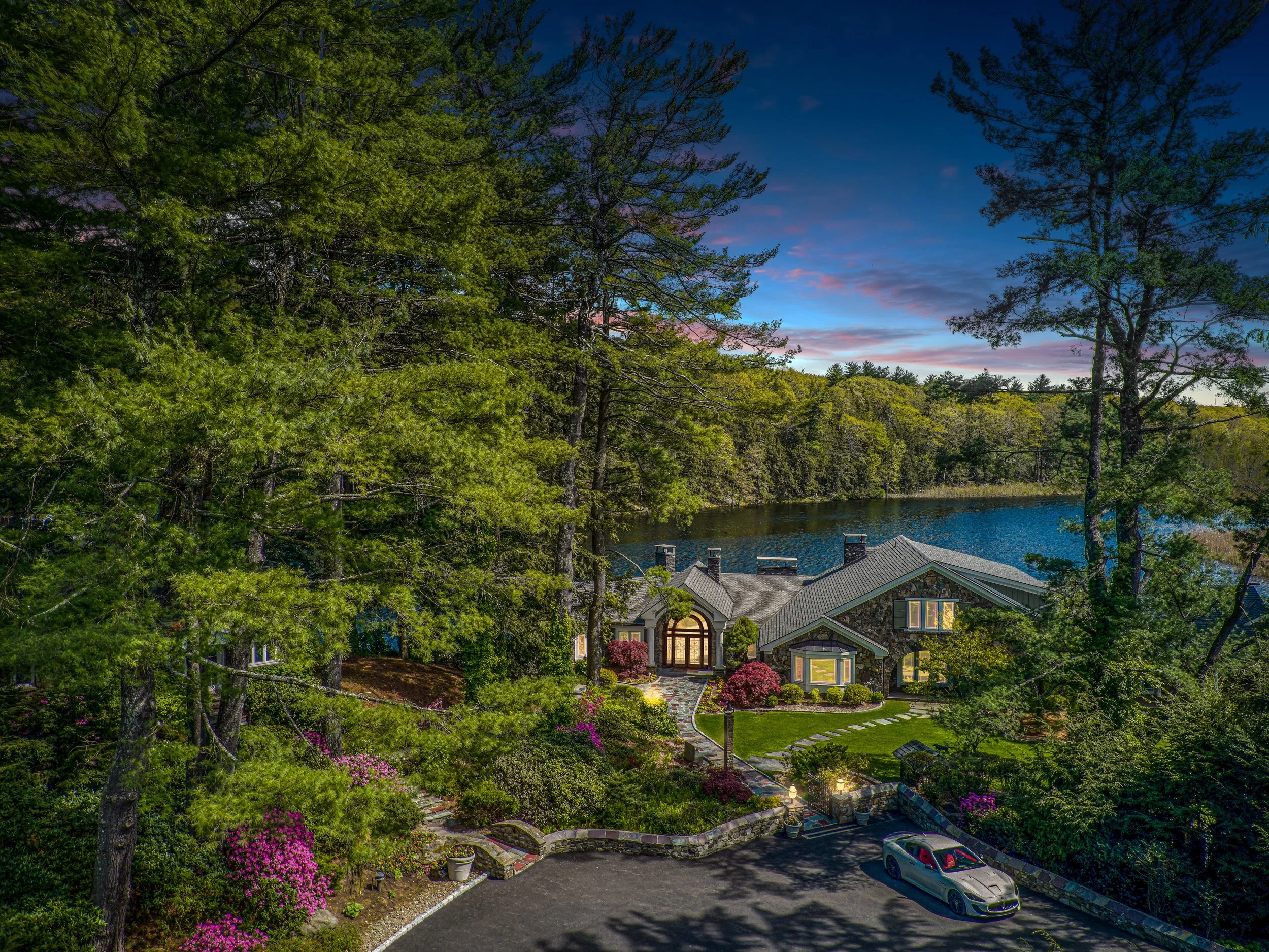 A large house with a stone exterior, front lawn, surrounded by trees and colorful bushes, near a lake, during dusk with a clear sky with pink and blue hues.