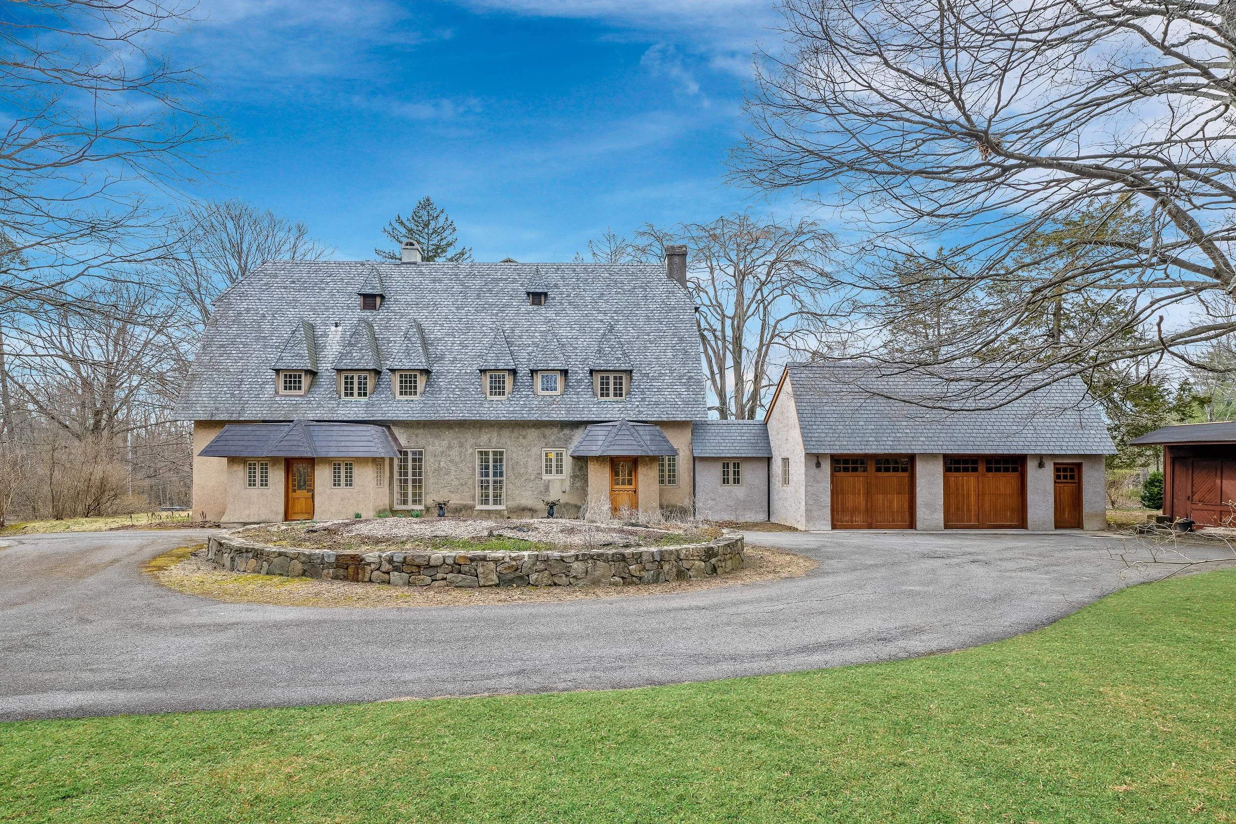 A large stone house with a gray shingled roof, multiple small dormer windows, and wooden doors, surrounded by trees and a circular driveway.