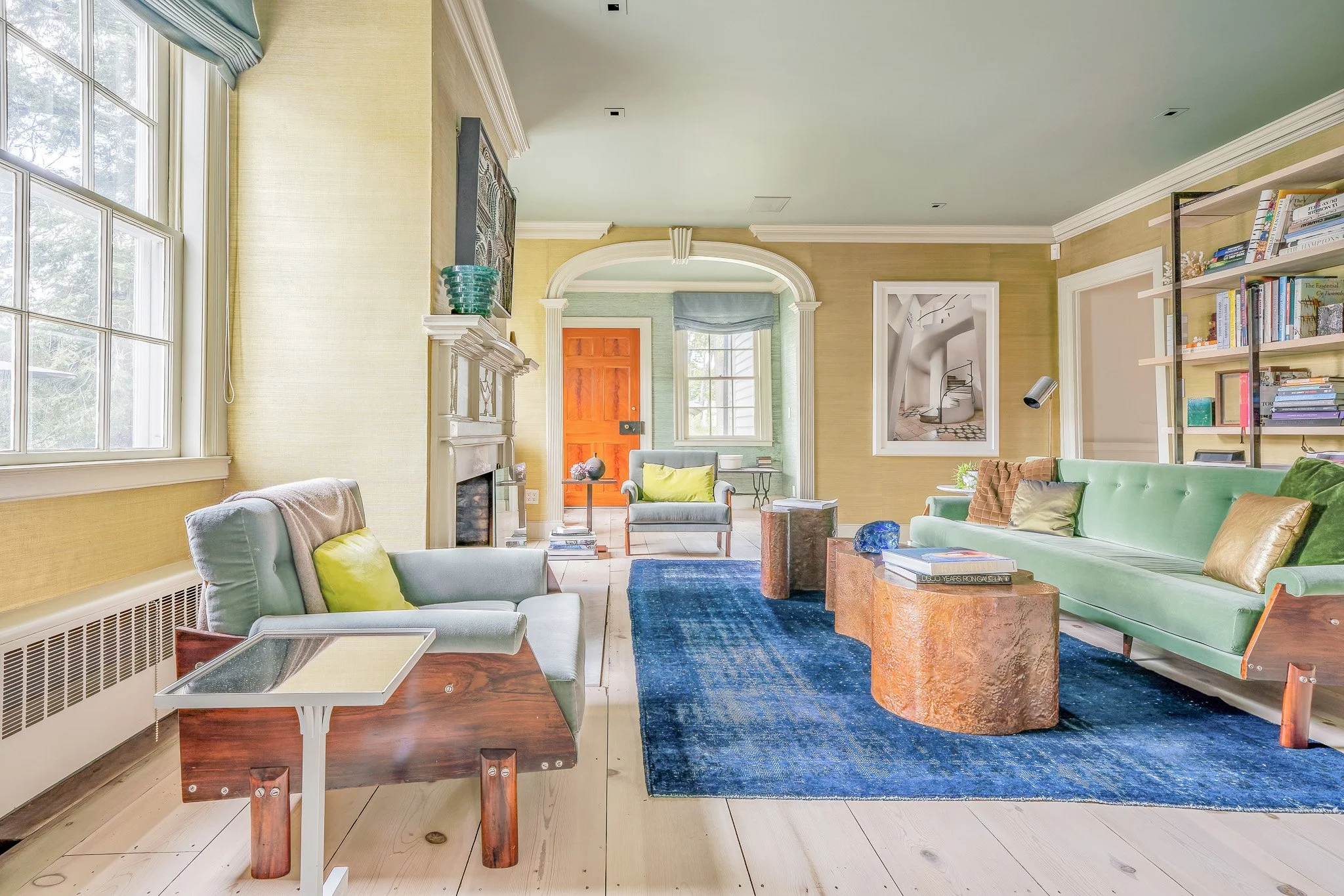Living room with light-colored walls, green sofa, vintage chairs, blue rug, wooden coffee tables, bookshelf, large windows, and decorative art pieces.