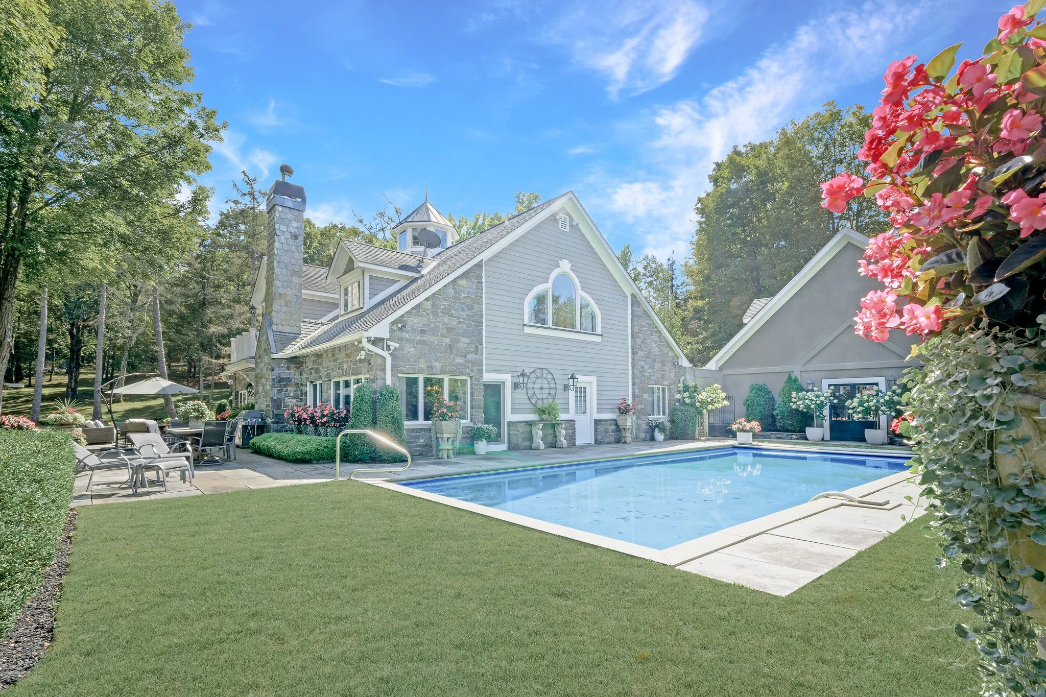 A backyard with a swimming pool, surrounded by greenery, with a large house featuring gray siding and stone accents, and outdoor furniture on a patio.