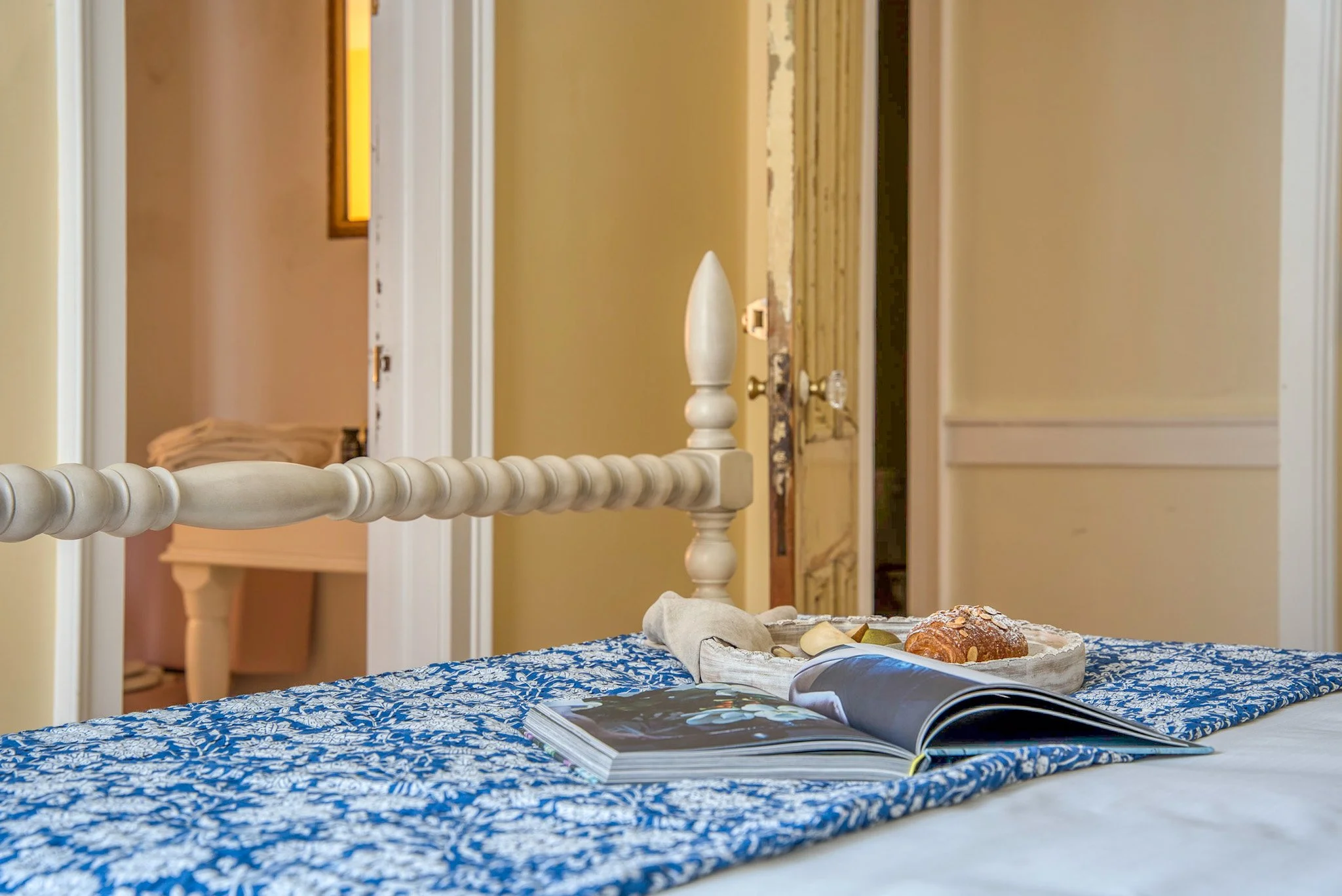 A bed with a blue and white patterned bedspread, an open magazine, a tray with a croissant and spoon, and a white bed rail, in a room with yellow walls and open doorway.