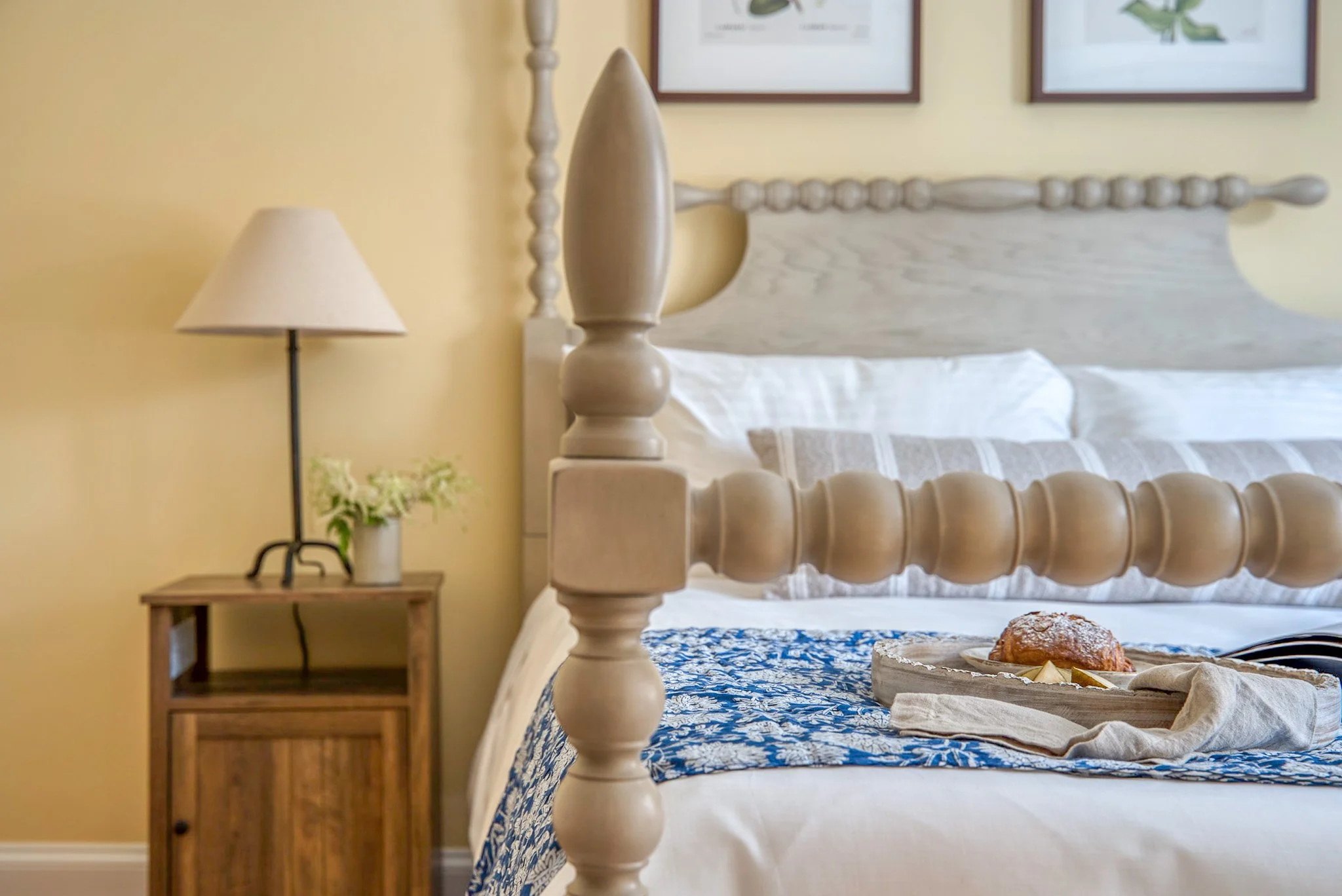 A bedroom with a wooden four-poster bed, a nightstand with a lamp, a small vase with flowers, and a tray with a pastry and butter on it.