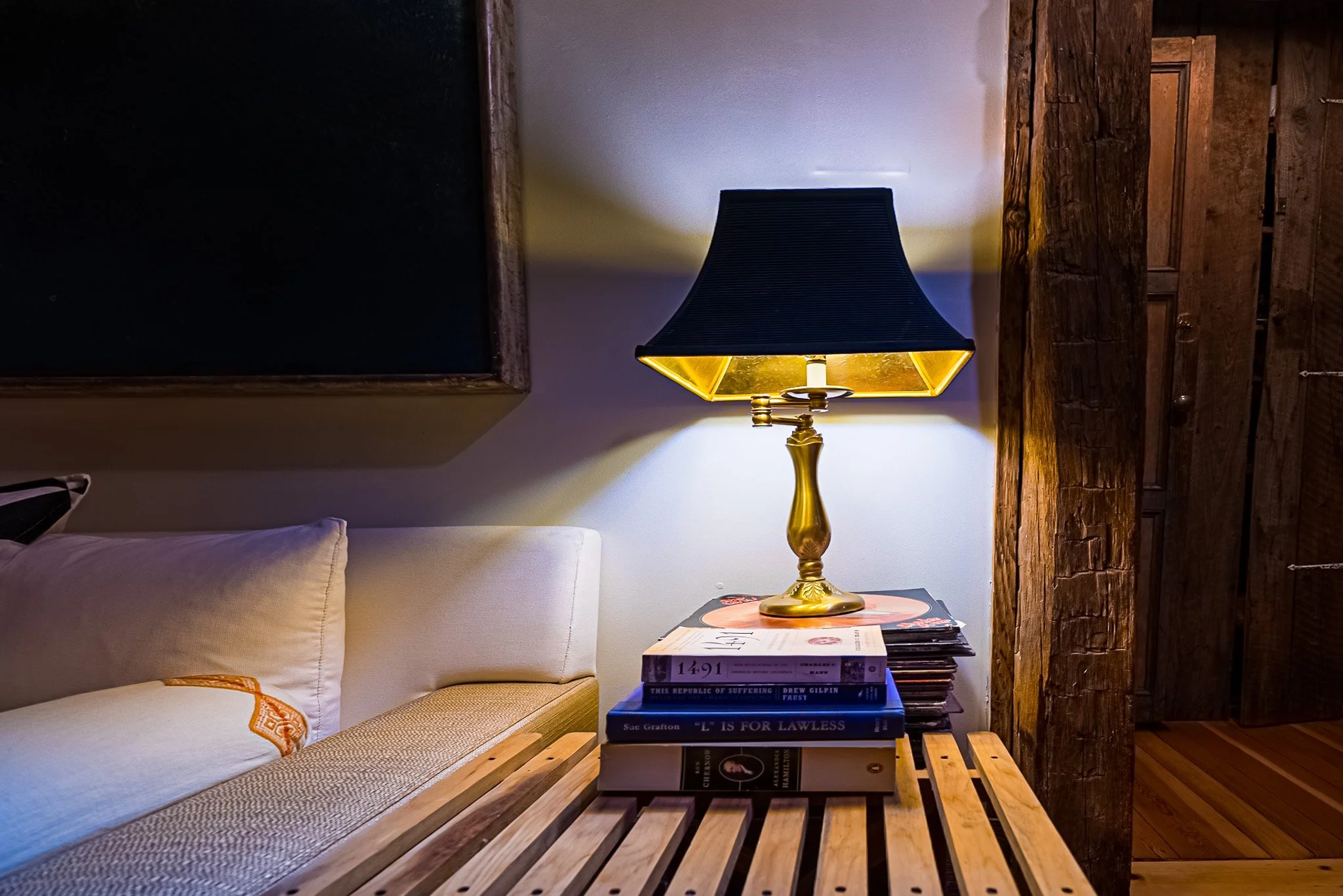 Side table with stacked books and a vintage lamp, next to a bed with white pillows and a black chalkboard, in a room with wooden structural beams.