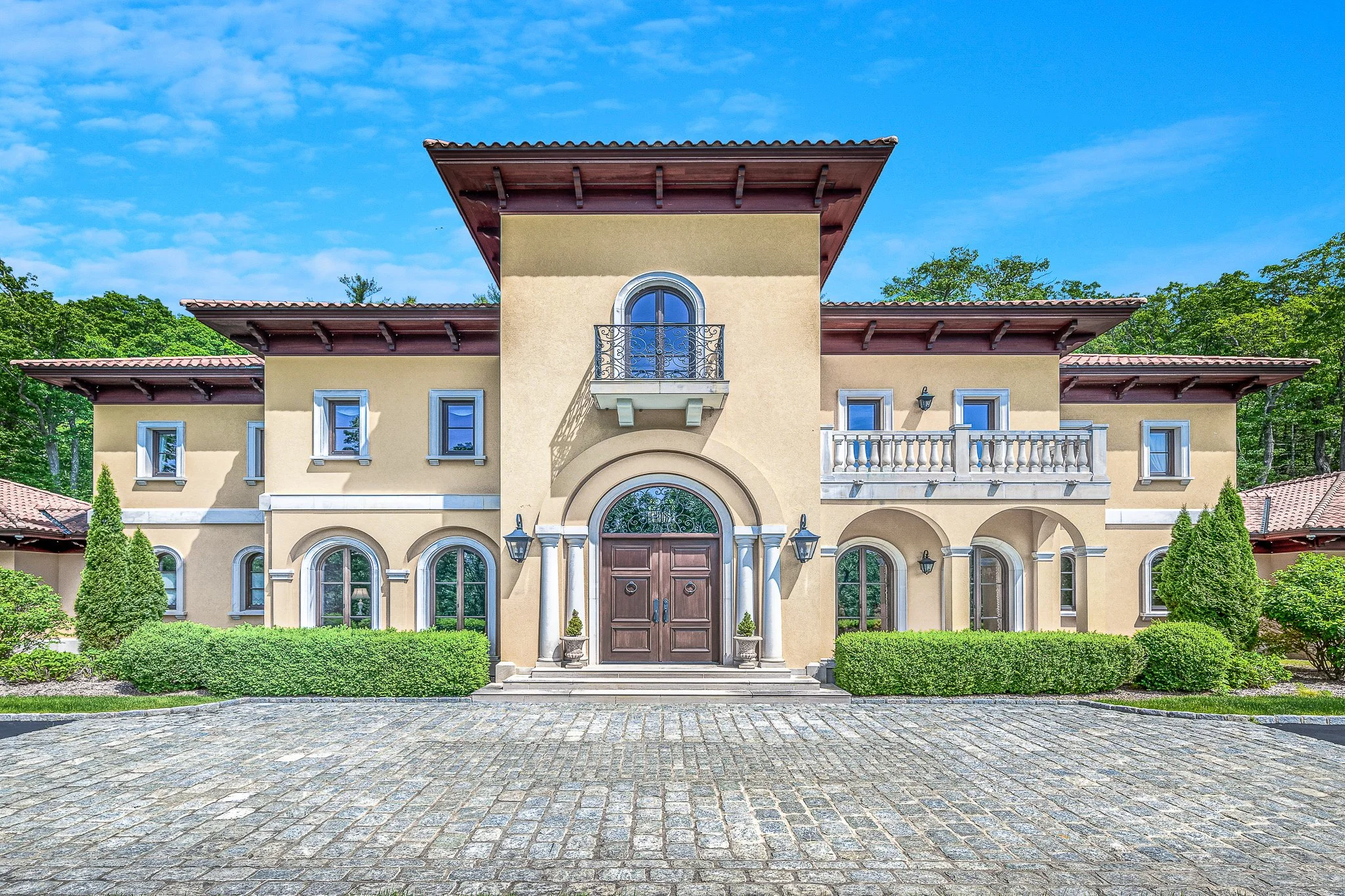 A large, elegant Mediterranean-style house with beige stucco walls, red tiled roof, arched windows, and a double front door, surrounded by green shrubbery and trees under a blue sky.