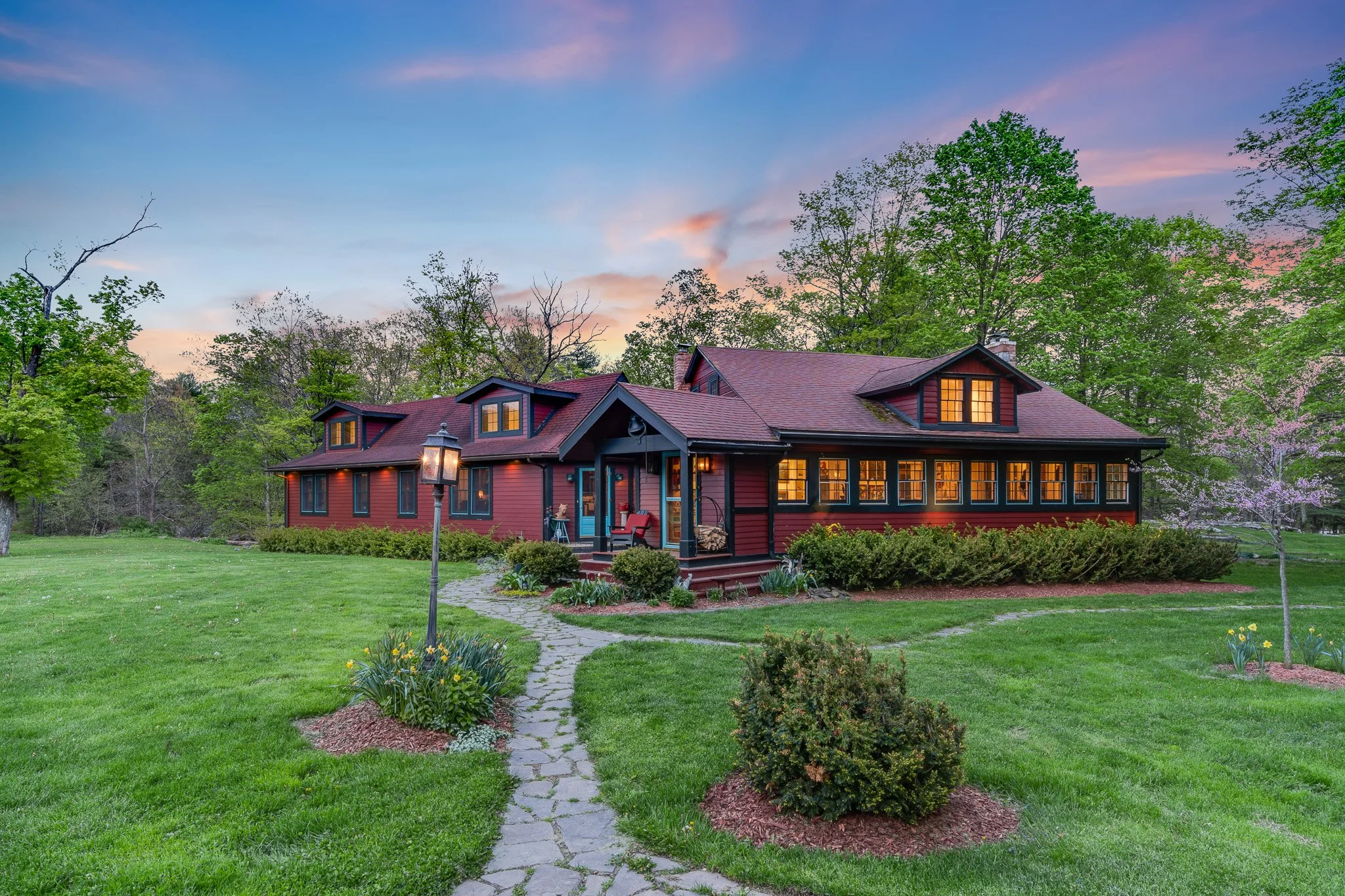 A large, red two-story house with black trim and glowing yellow windows, set amidst green lawns and trees at dusk, with a stone pathway leading to the front porch.