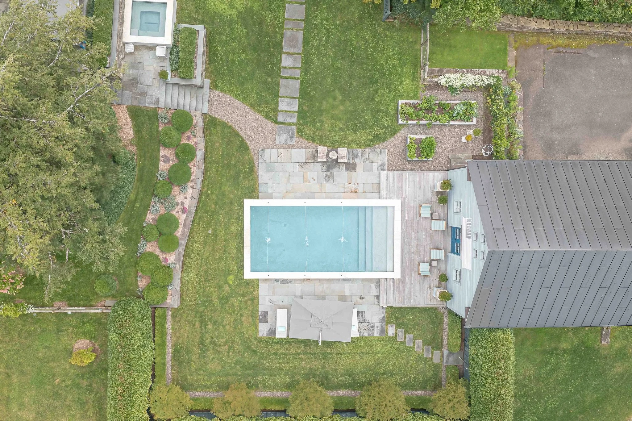 Aerial view of a backyard with a rectangular swimming pool, patio area with chairs, well-maintained lawn, garden beds, and surrounding trees and shrubs.