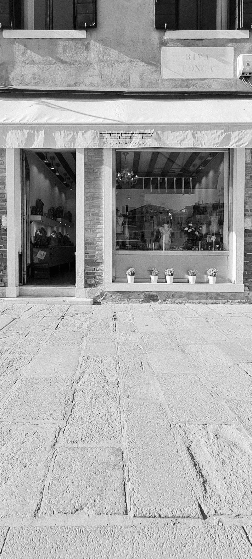 Esterno di un negozio con vetrina e piante in vaso, scritto 'Riva Love' su una tenda sopra l'ingresso, con vasi e piante visibili all'interno.