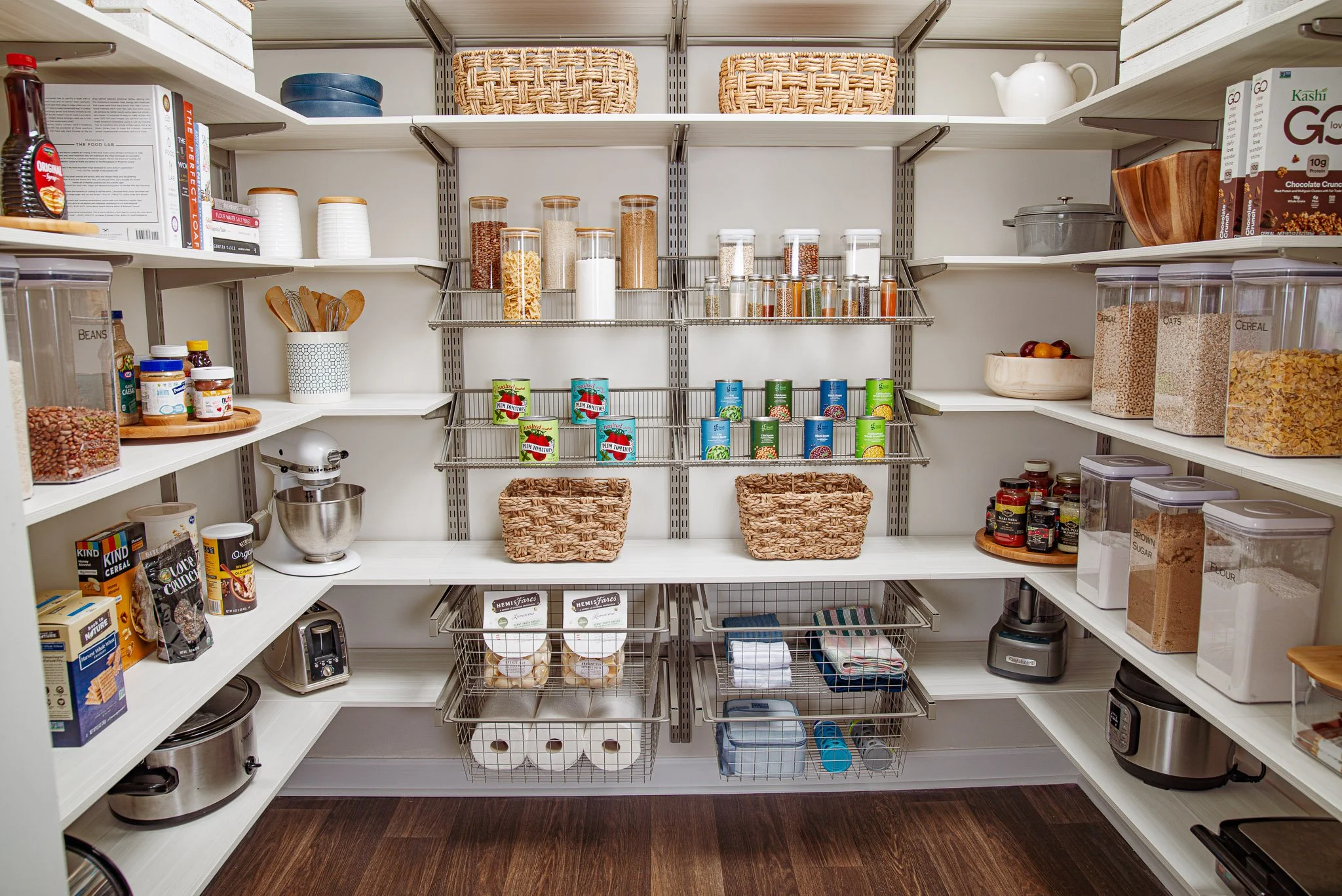 Organized walk-in pantry with adjustable shelves and basket-style drawers containing canned goods, cereals, jars, and kitchen appliances