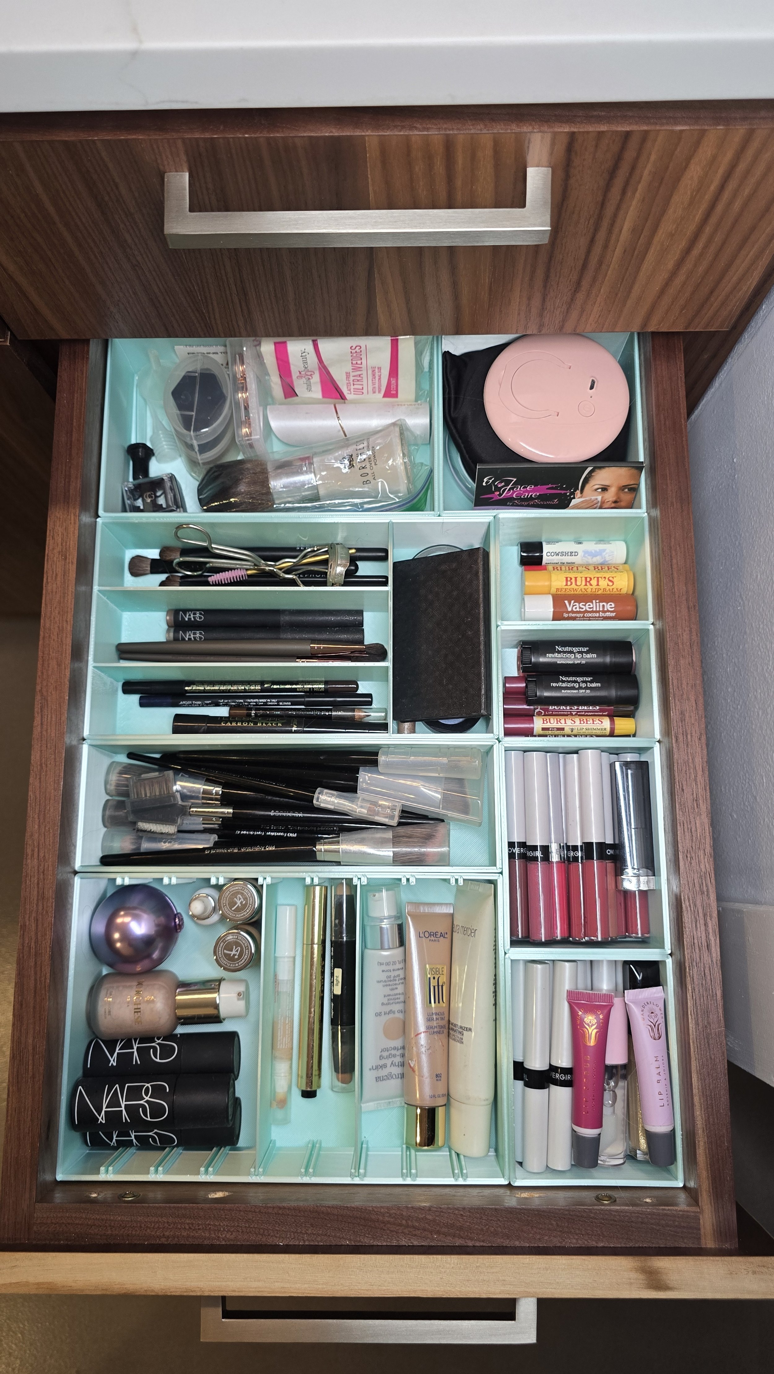 Open wooden drawer containing various makeup and skincare products, brushes, lip balms, and cosmetics organized in compartments.