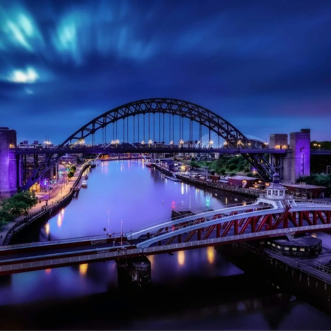 Nighttime cityscape of a river with illuminated bridges and city lights, cloudy sky overhead.