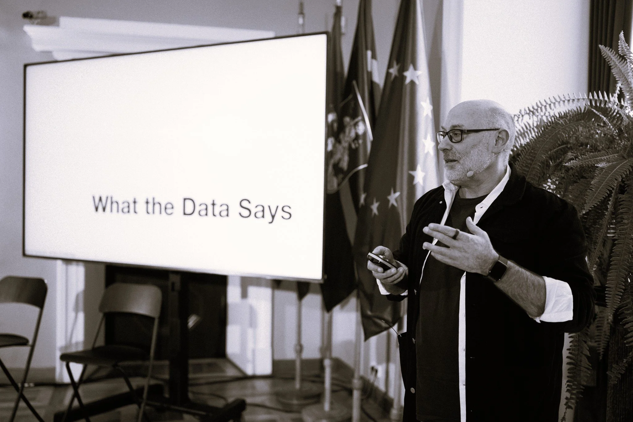 A man with glasses, wearing a face microphone, stands next to a large screen that displays the text 'What the Data Says.' He is holding a phone and gesturing with his other hand during a presentation indoors, with flags and plants in the background.