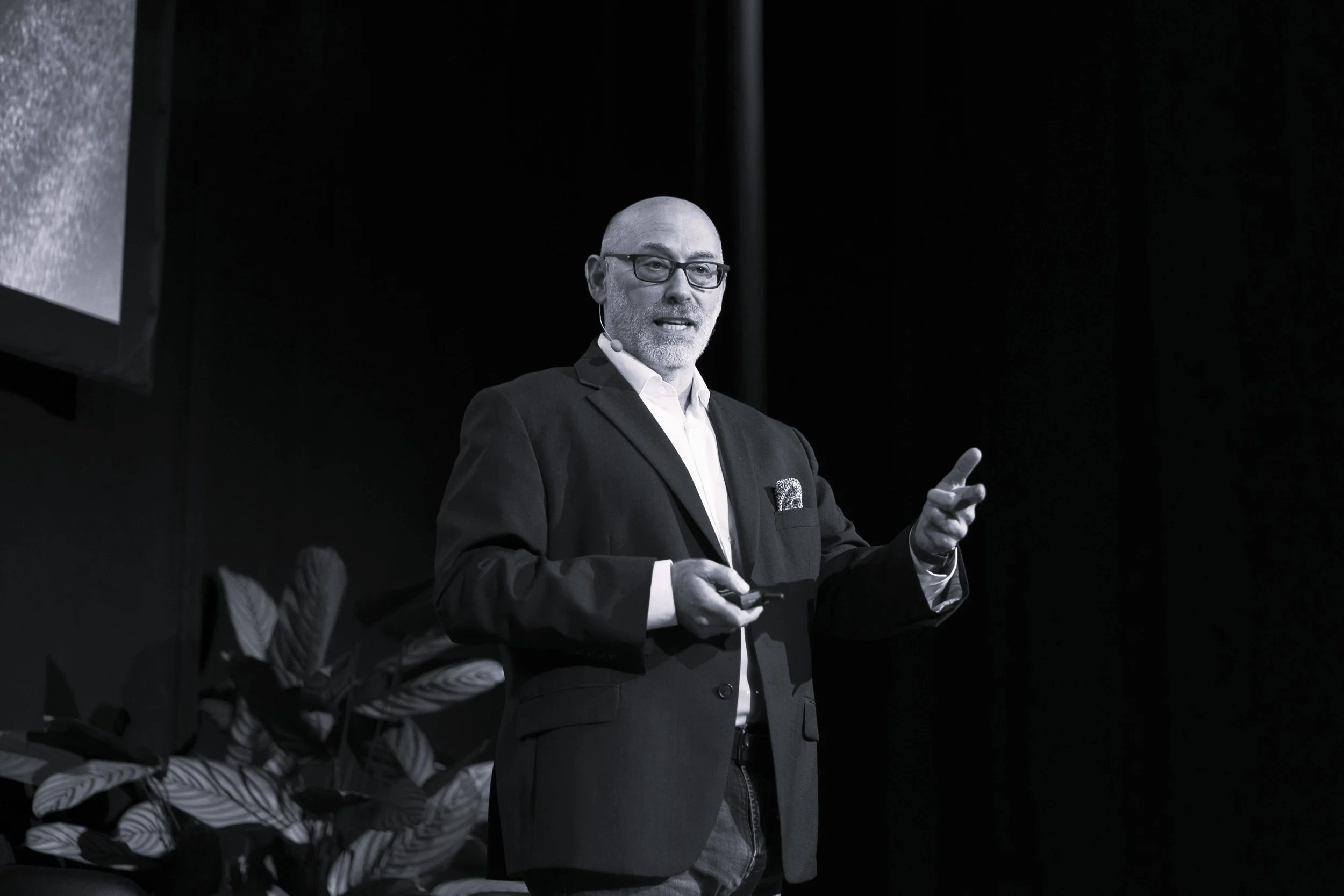 A man in a suit and glasses giving a presentation on stage, with a microphone headset, in front of a dark background with potted plants.