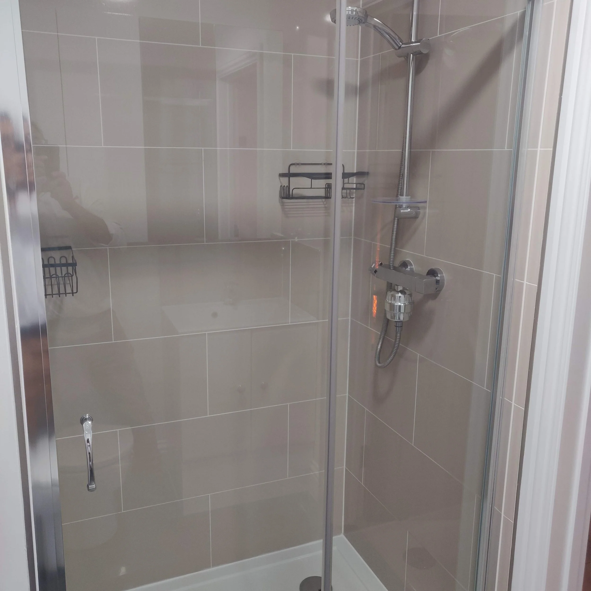 Empty glass shower enclosure with beige tiled walls and a silver showerhead, shelf, and control handle.