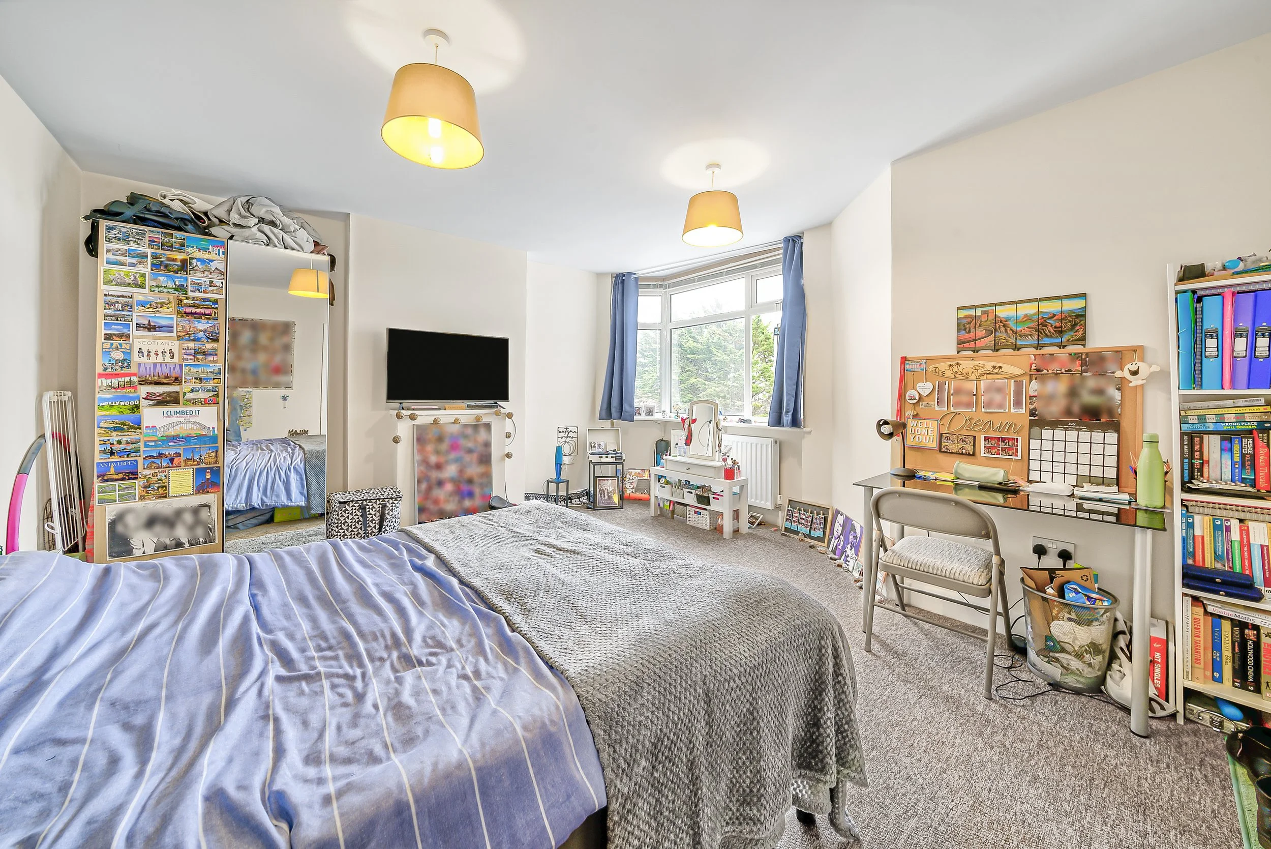 A bedroom with a bed covered in a gray blanket and striped sheets, a window with blue curtains, a TV on the wall, shelves with books and decorations, and a mirror.