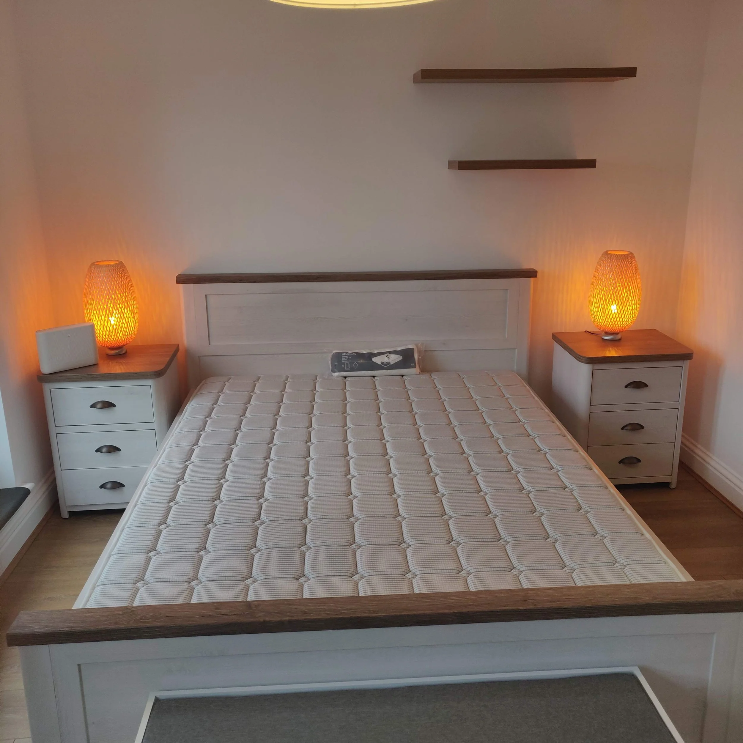 Bedroom with a white bed, two white nightstands with wooden tops, each with a orange woven table lamp, two floating wooden shelves on the wall above the bed, a small decorative pillow on the bed, and wood flooring.