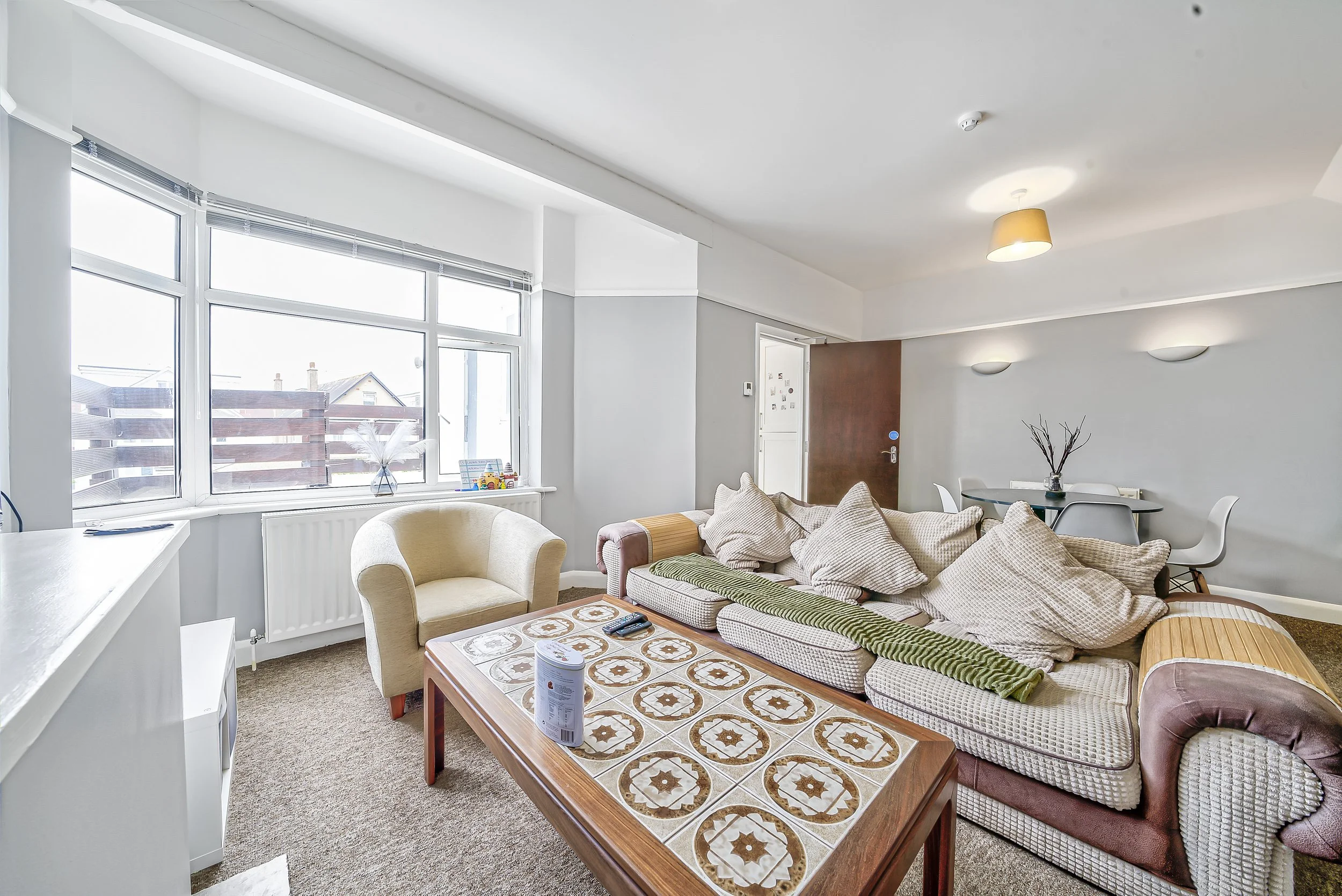 Living room with large window, beige armchair, patterned coffee table, sofa with multiple pillows, dining table with chairs, and minimal decor.