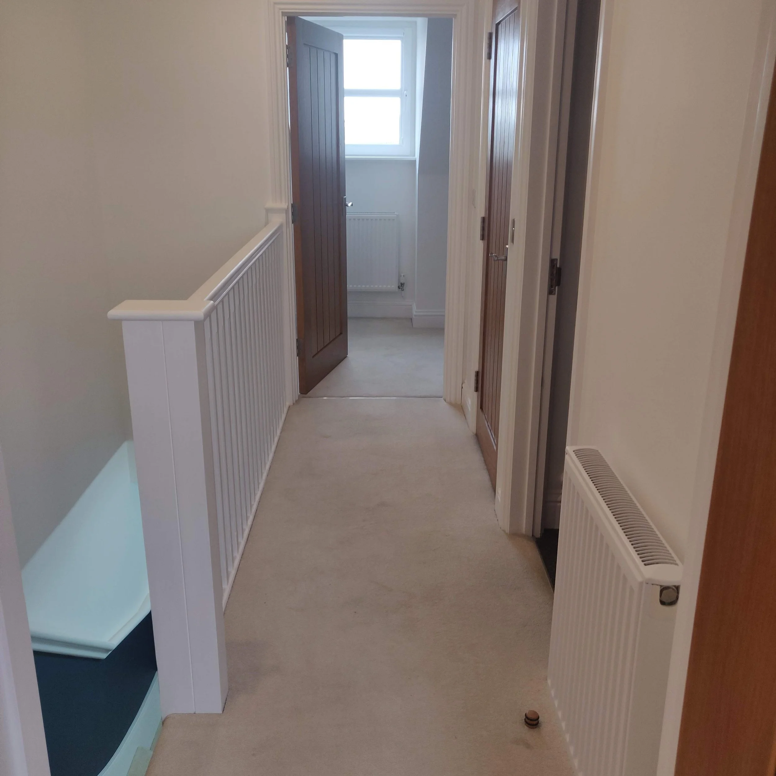 Corridor leading to a door with a window, with white walls, beige carpet, and a white radiator on the right side.