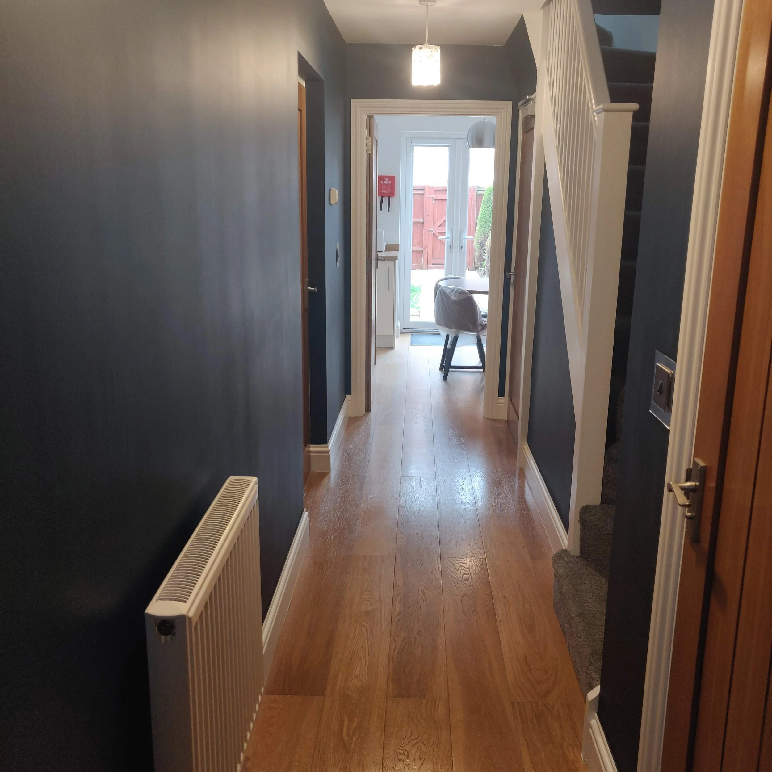 A hallway in a house with wooden flooring, dark blue walls, a staircase with a white railing on the right, a radiator on the left, leading to a dining area with a glass door at the far end, allowing natural light inside.