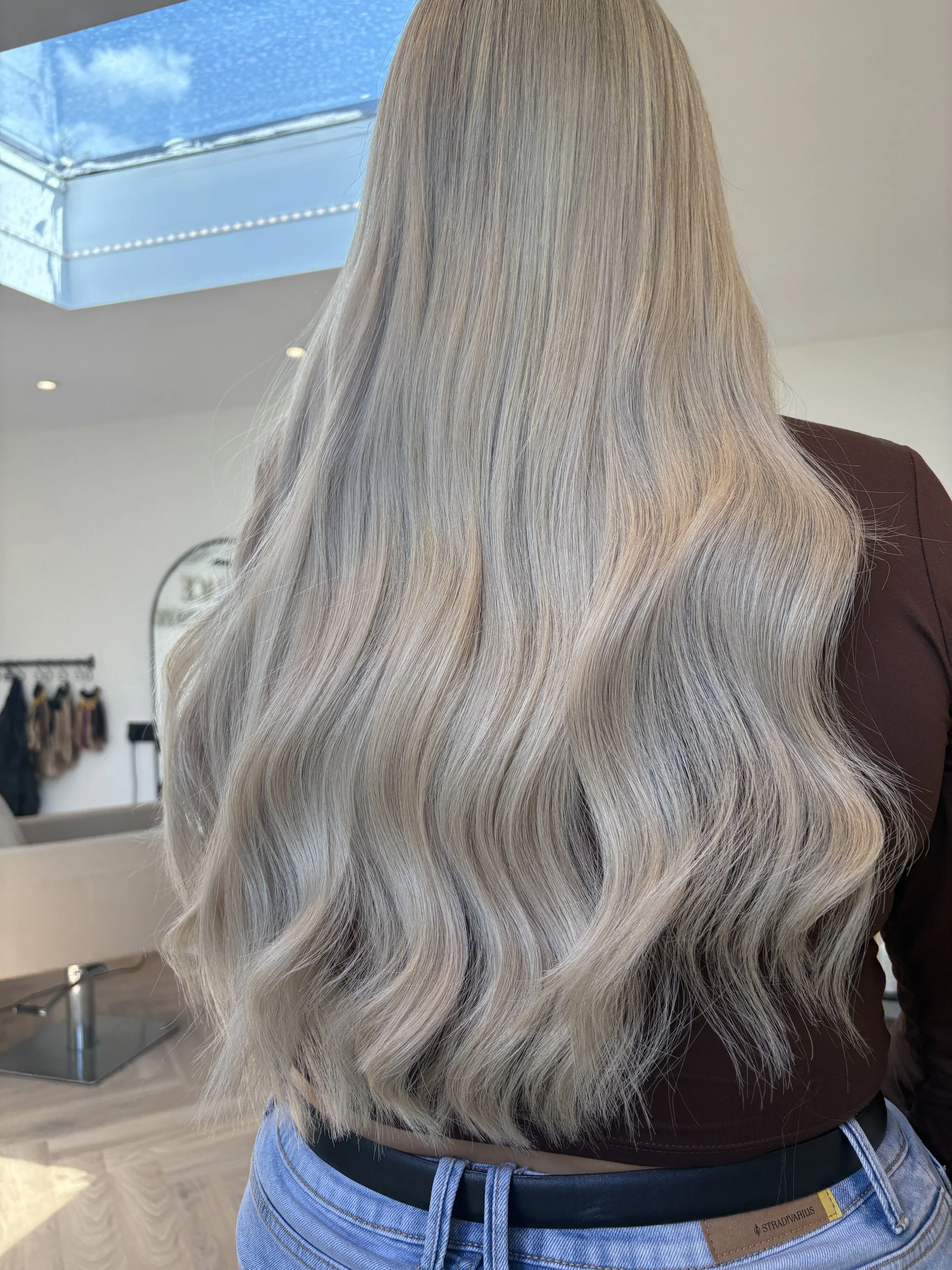A person with long, wavy ash blonde hair extensions, in a modern minimal training salon.