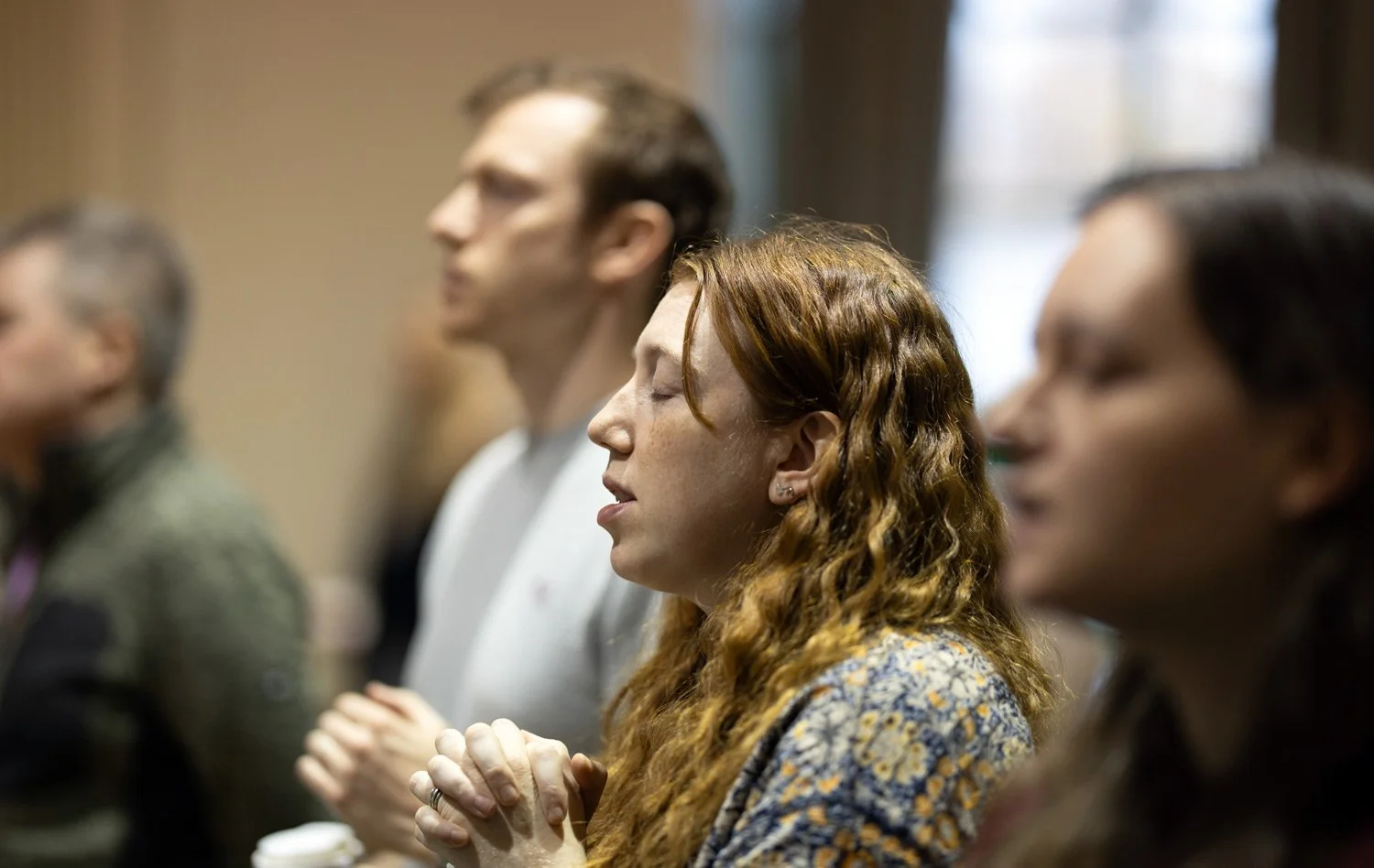 People sitting in a row with eyes closed, hands clasped, appearing to pray or meditate in a peaceful setting.