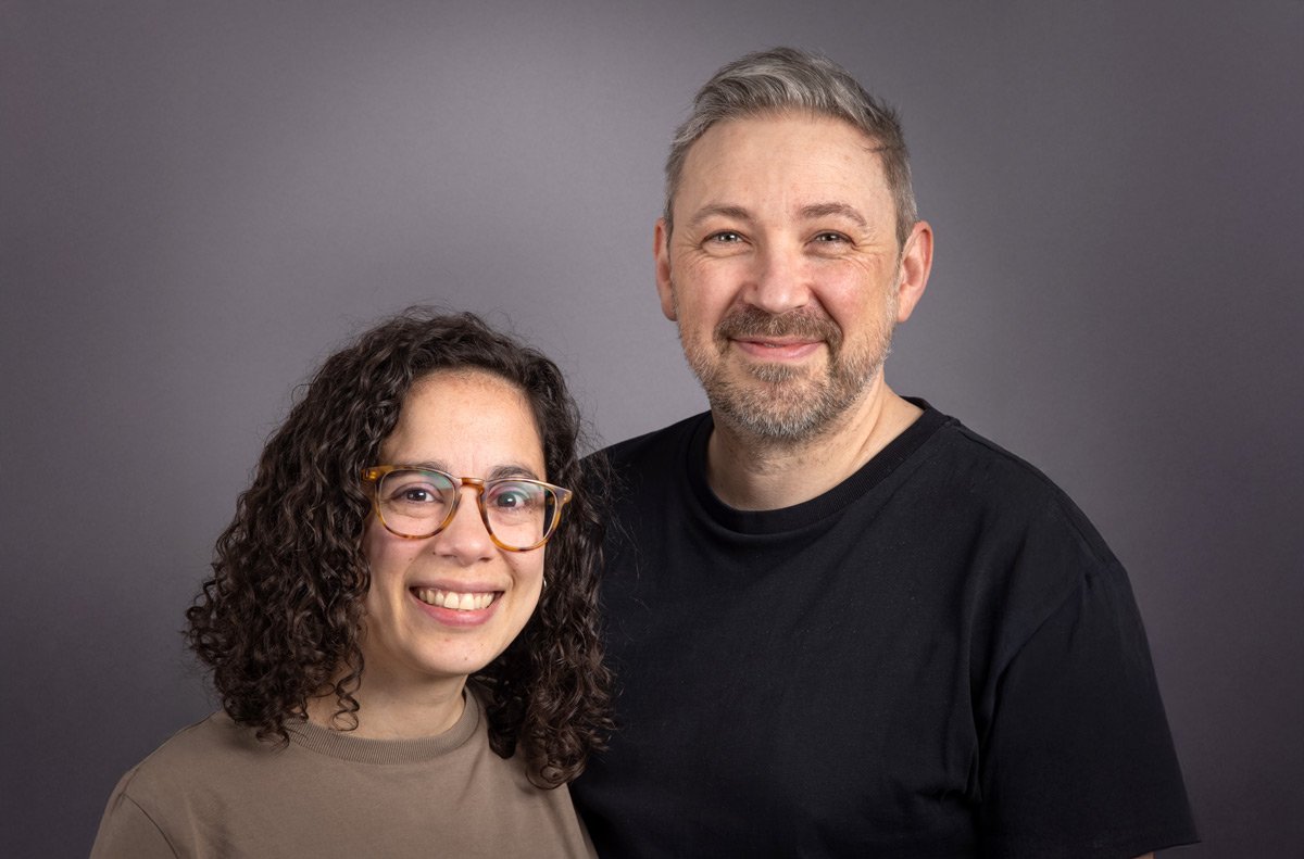 A smiling woman with curly dark hair and glasses standing next to a smiling man with gray hair and beard. Both are wearing casual clothing and are posing in front of a plain gray background.