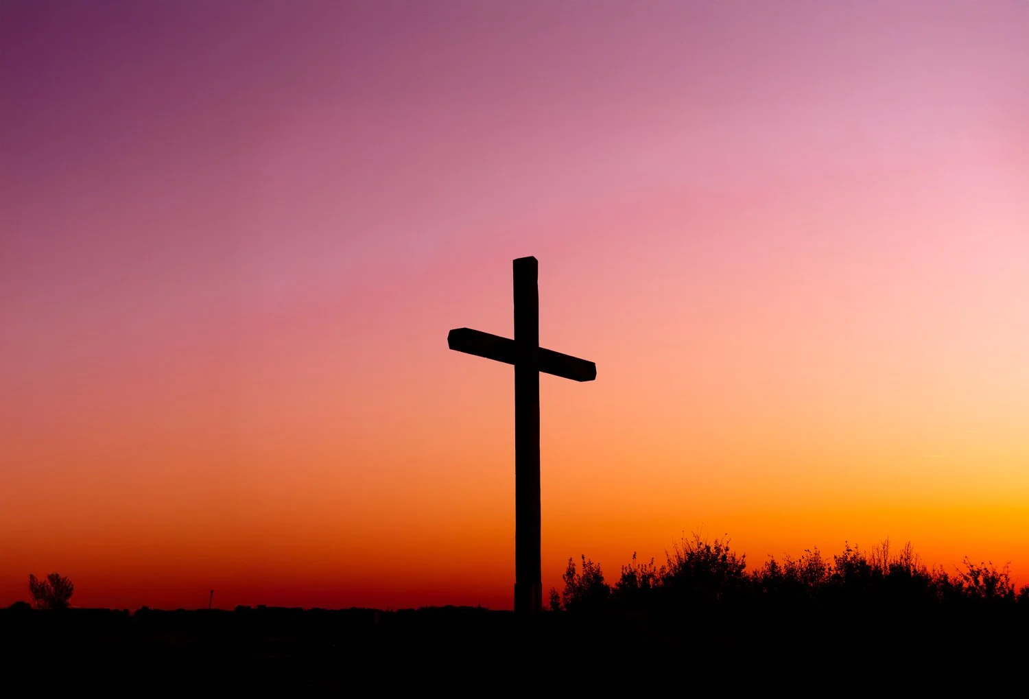 Silhouette of a cross against a colorful sunset sky with orange, pink, purple hues and dark bushes in the foreground.