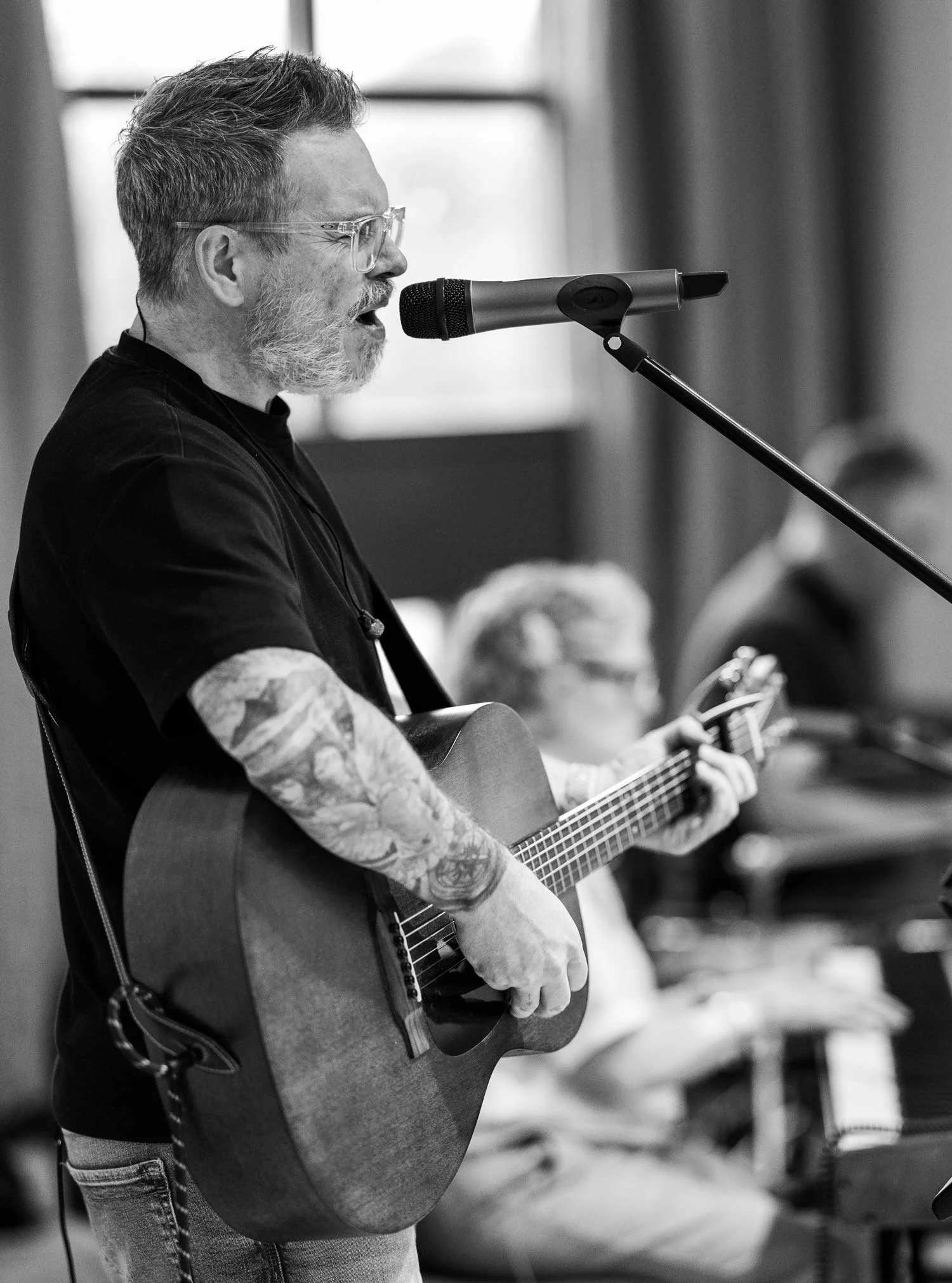 A man with glasses and a beard, wearing a black T-shirt with tattoos on his arms, playing an acoustic guitar and singing into a microphone during a performance in an indoor setting.