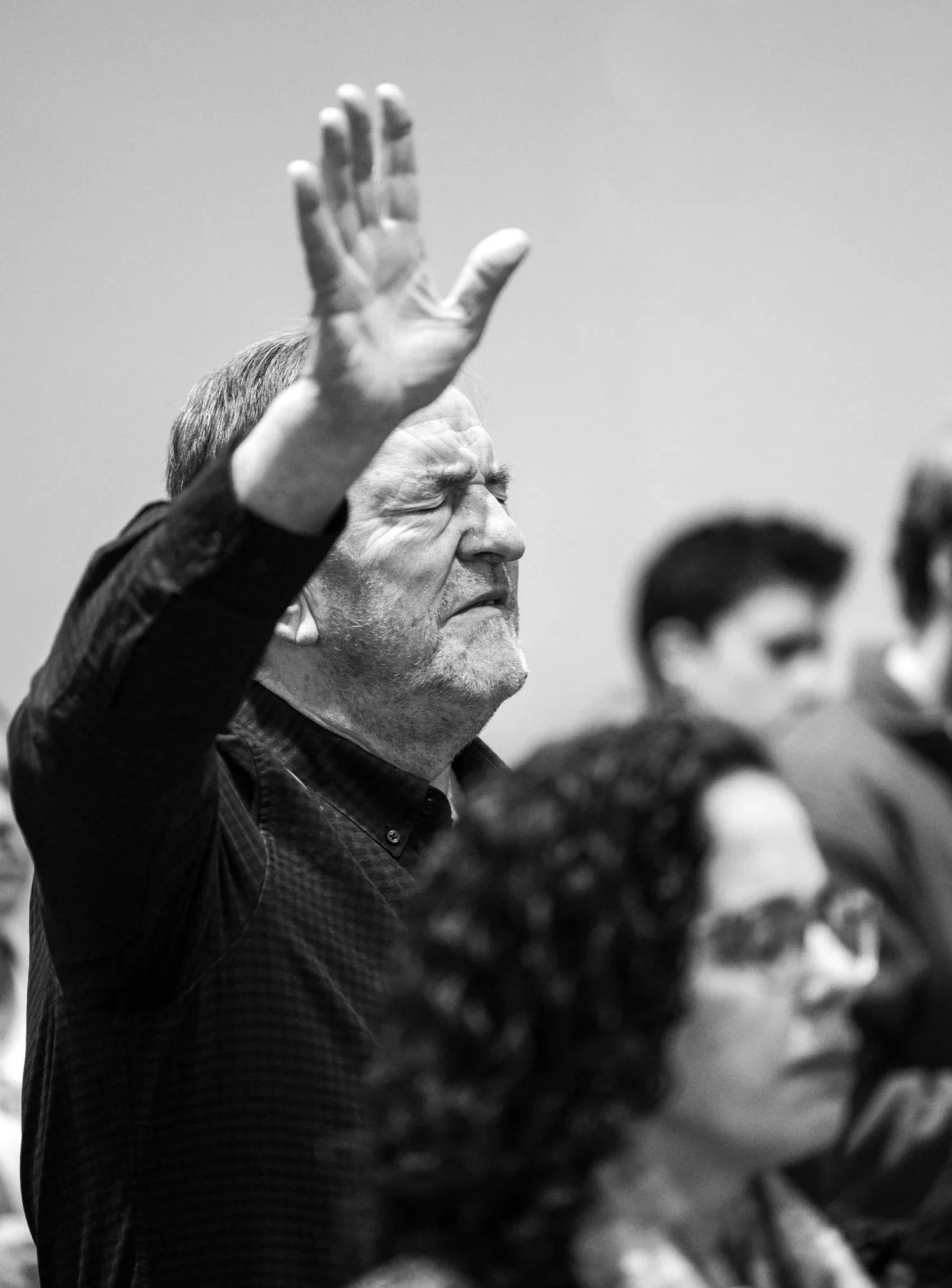 A black-and-white photo of a man with closed eyes, raising his right hand, possibly in a prayer or worship setting, with several other people visible in the background.