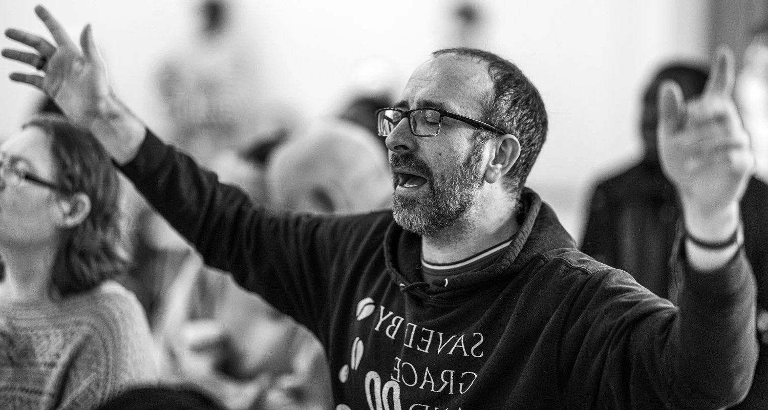 Man with glasses and beard with eyes closed, raising his arms, praying or singing, surrounded by a group of people.