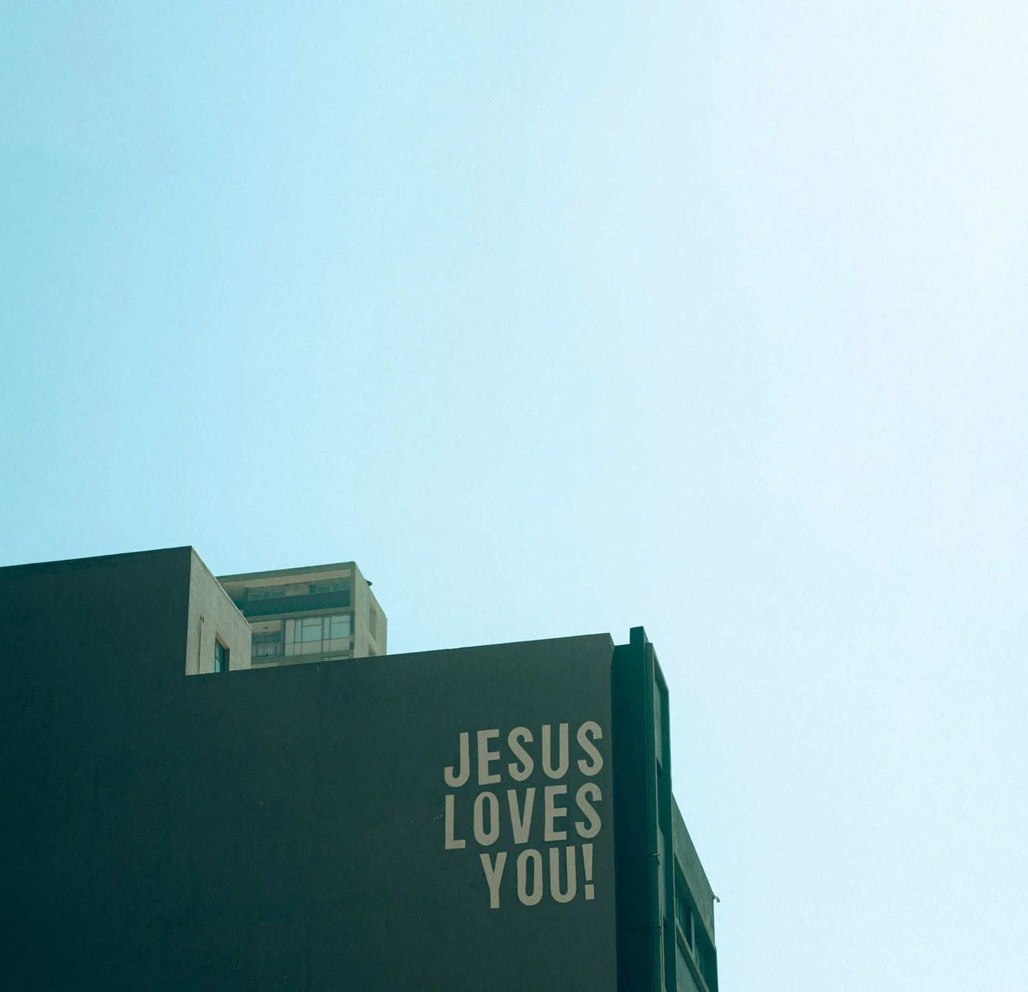 A building with a sign that reads 'JESUS LOVES YOU!' against a clear blue sky.