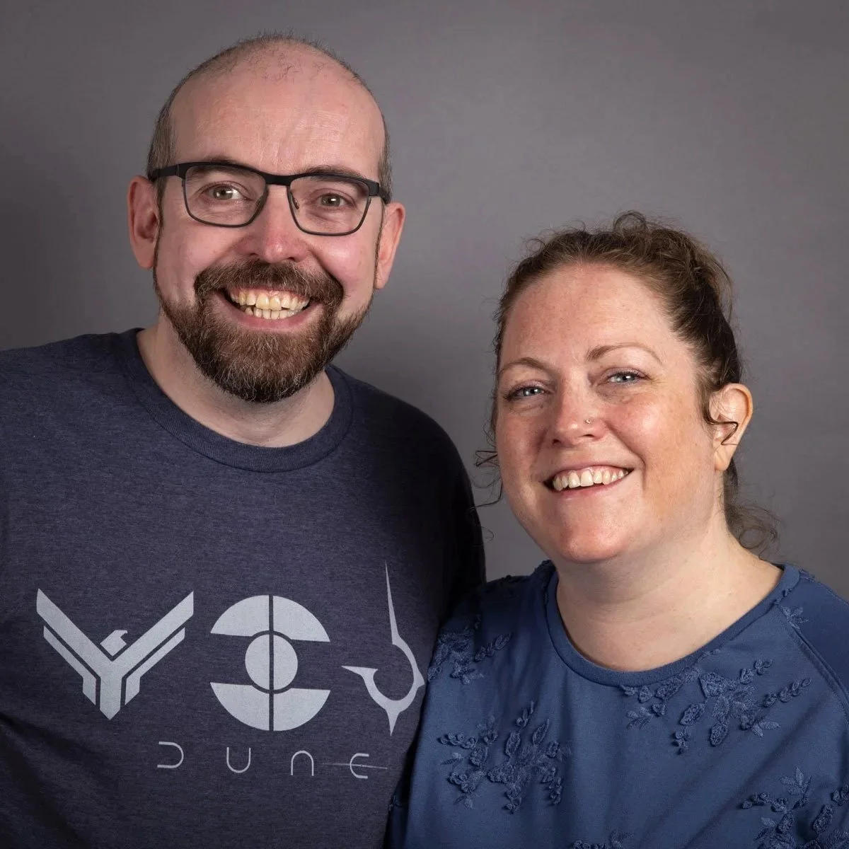 A smiling man with glasses and a beard wearing a dark blue t-shirt, and a smiling woman with curly hair in a blue shirt, standing together against a gray background.