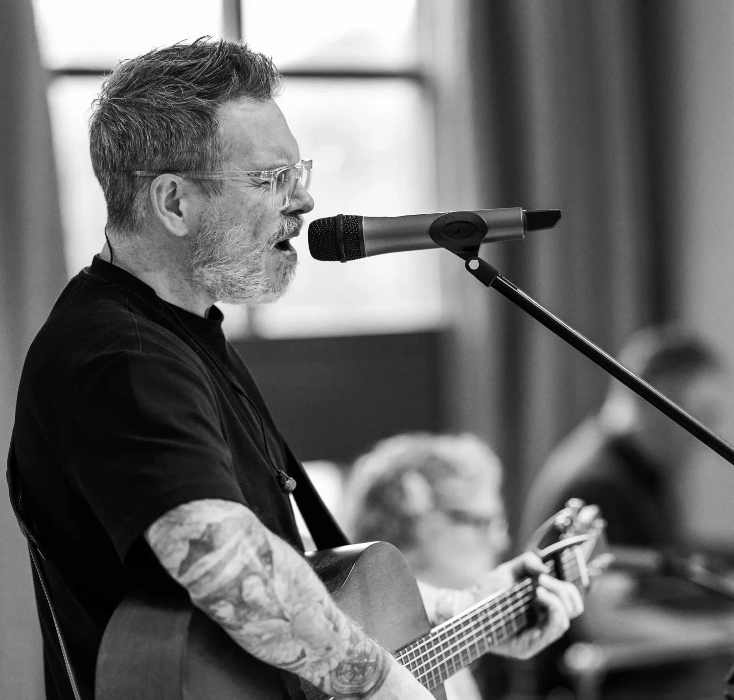 A man with glasses and a beard, wearing a black T-shirt with tattoos on his arms, playing an acoustic guitar, leading worship at Falkirk Vineyard Church.