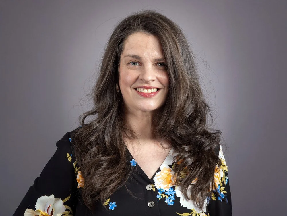 A woman with long wavy brown hair smiling, wearing a black floral blouse, against a gray background.