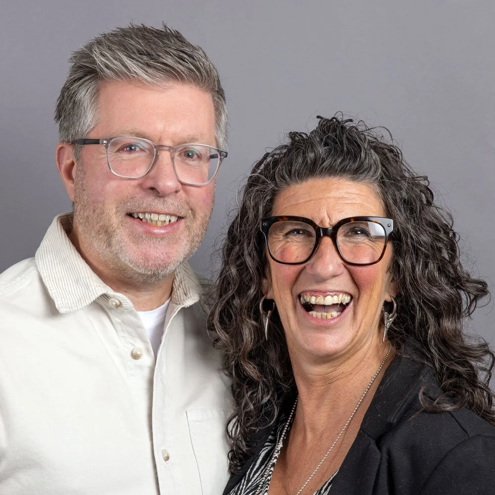 A smiling man with glasses and a woman with curly hair and glasses, both wearing casual clothing, posing together against a gray background.