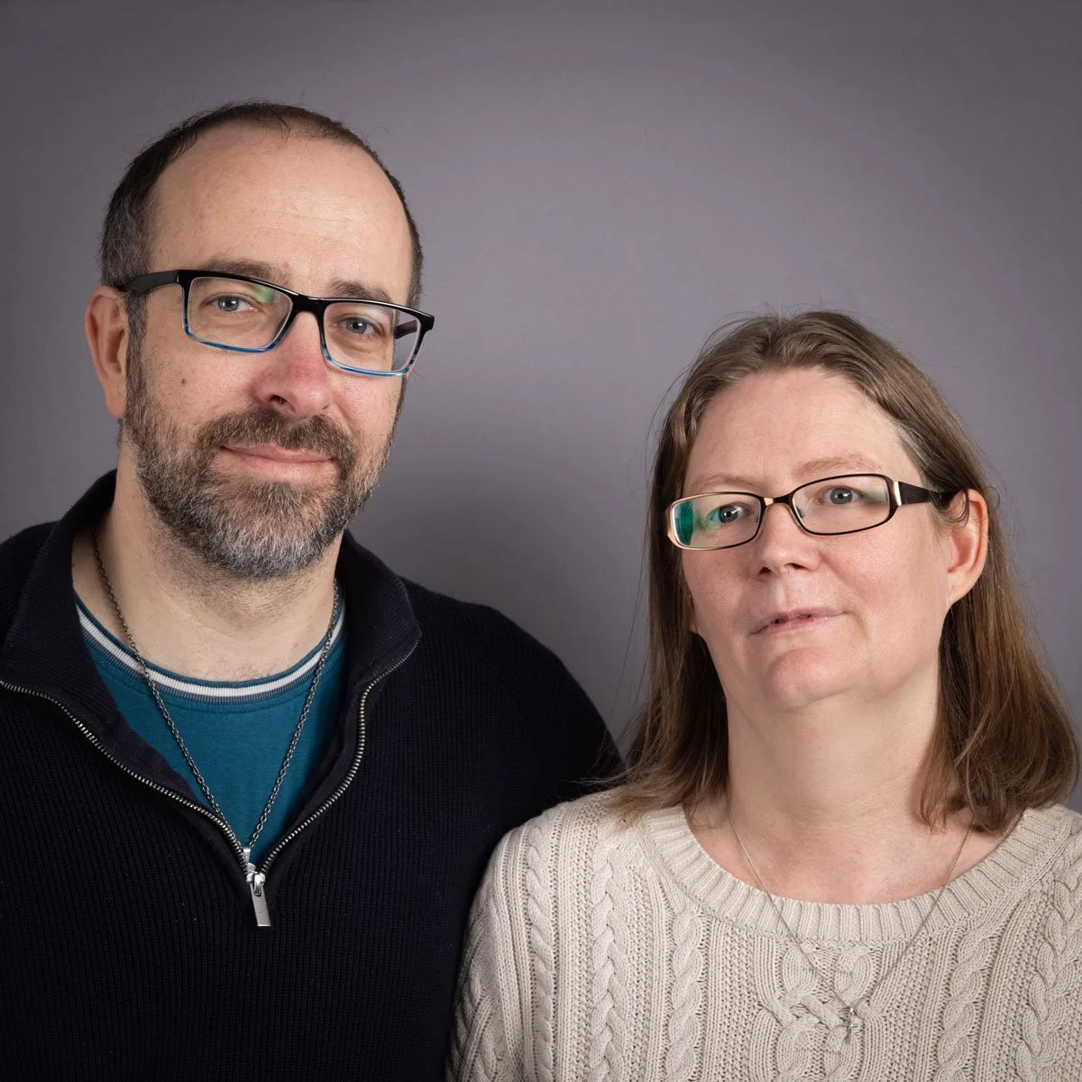 A middle-aged man with glasses and a beard standing next to a middle-aged woman with glasses and shoulder-length hair against a gray background.