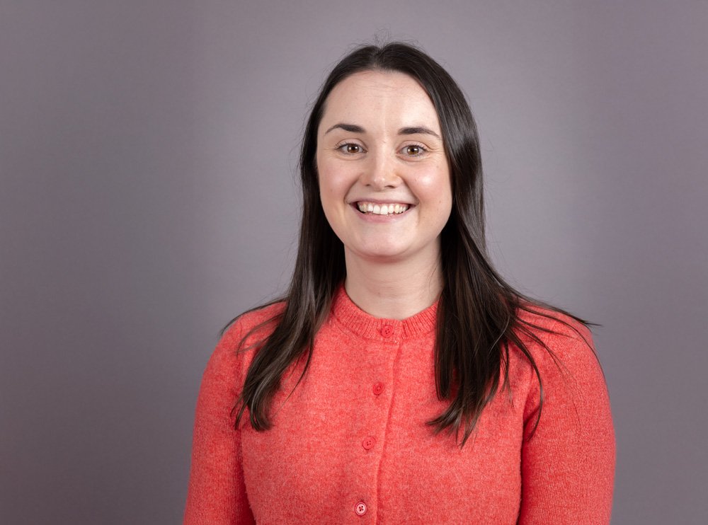 A woman with long dark hair wearing a red sweater, smiling against a gray background.
