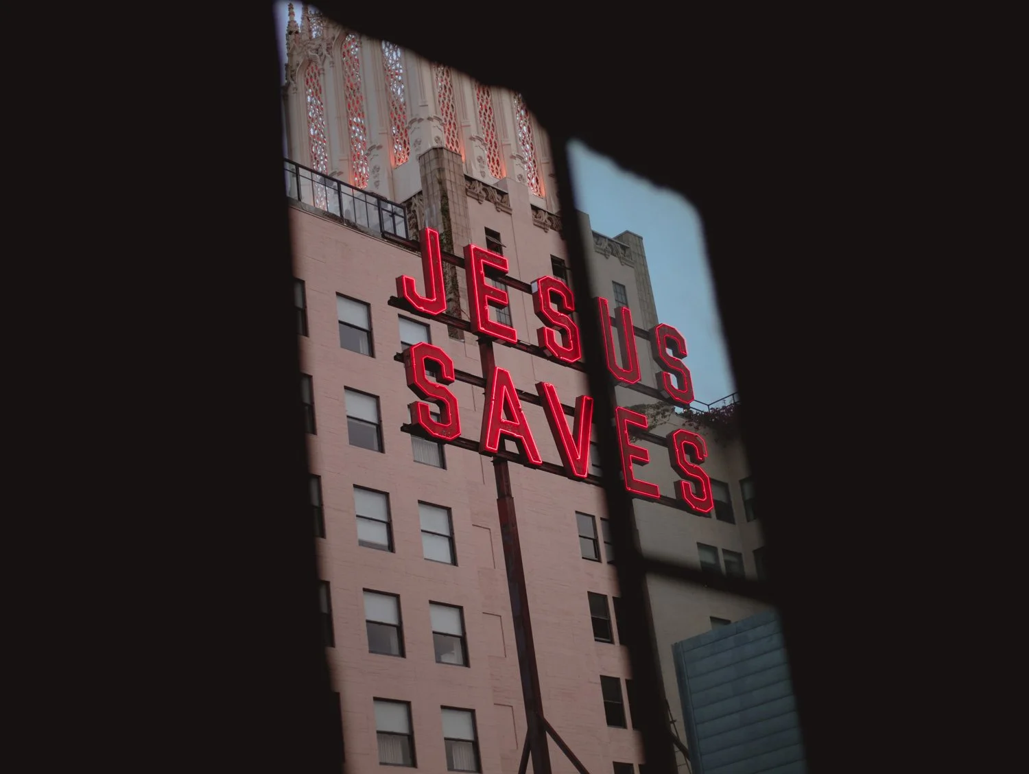 Neon sign reading 'Jesus Saves' on a building viewed through a window.
