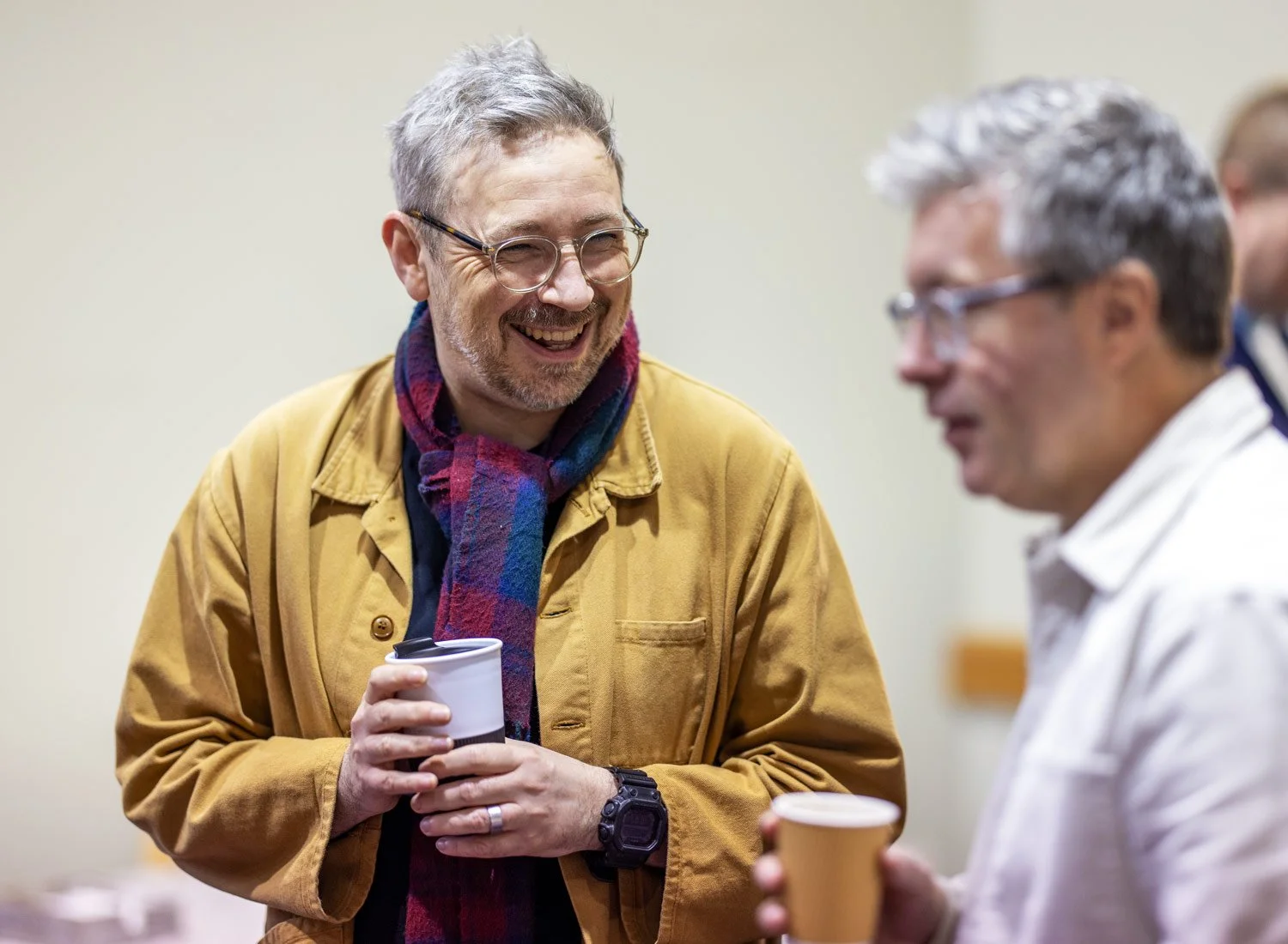 Two middle-aged men with gray hair and glasses are talking and smiling at each other while holding coffee cups.