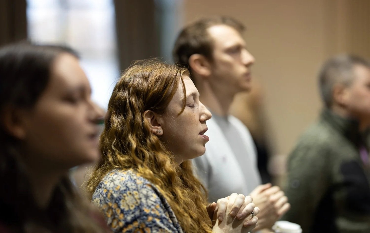 People meditating with eyes closed, sitting in a row, in a room with warm lighting.