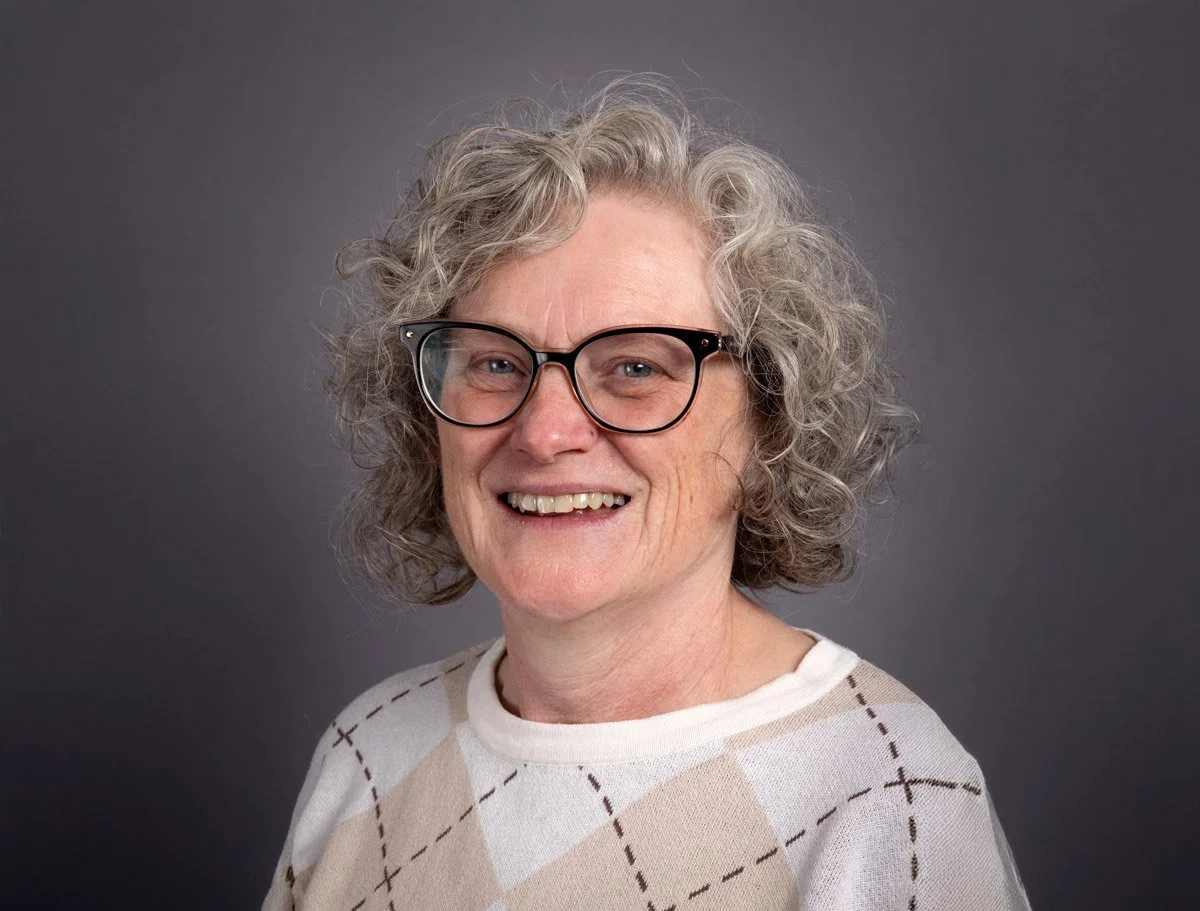 Portrait of an older woman with curly gray hair, wearing glasses and a beige and white argyle sweater, smiling against a dark gray background.