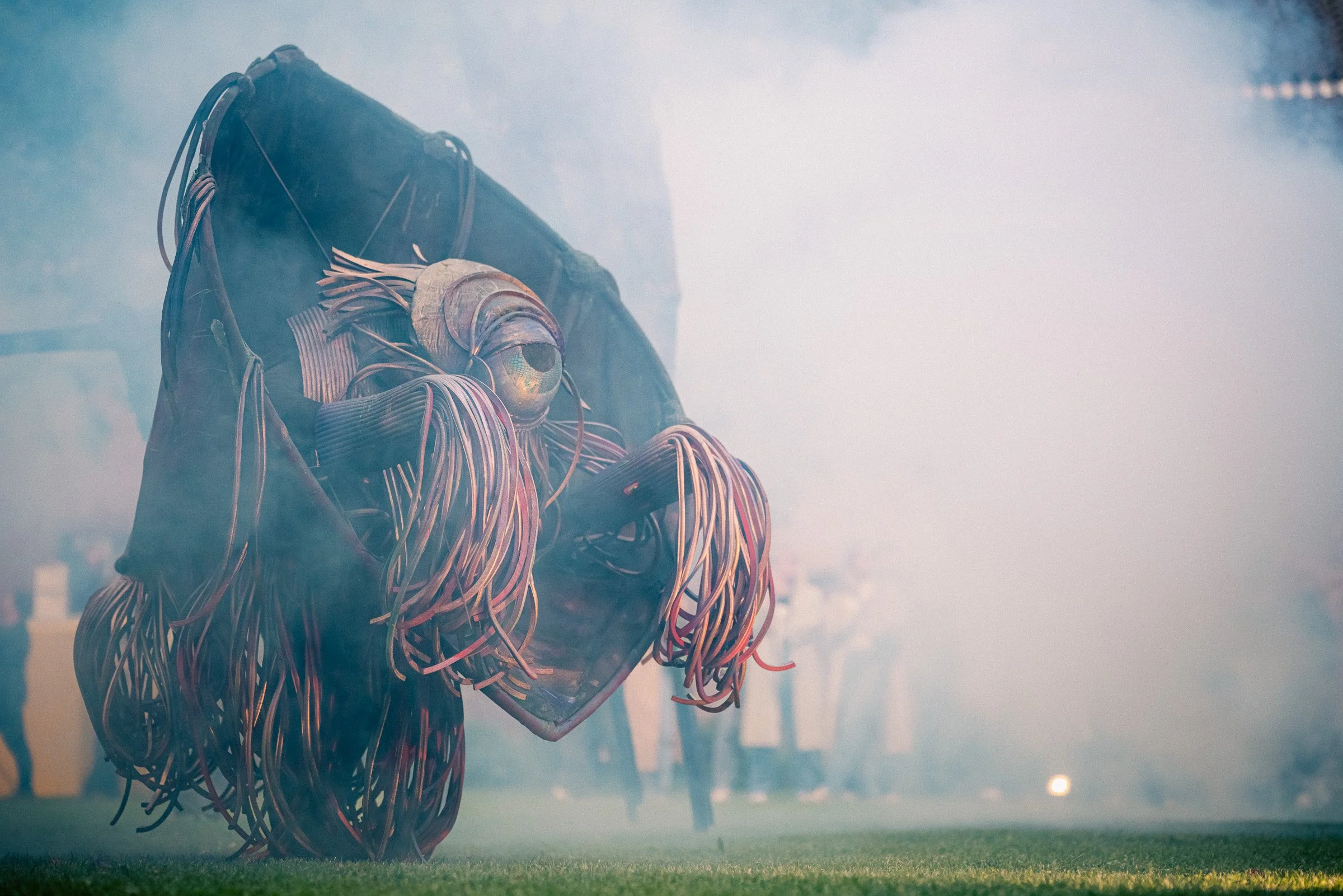 A futuristic sculpture of a humanoid figure with a large eye for a head, covered in tangled wires, situated outdoors with smoke and fog, and people in the background.