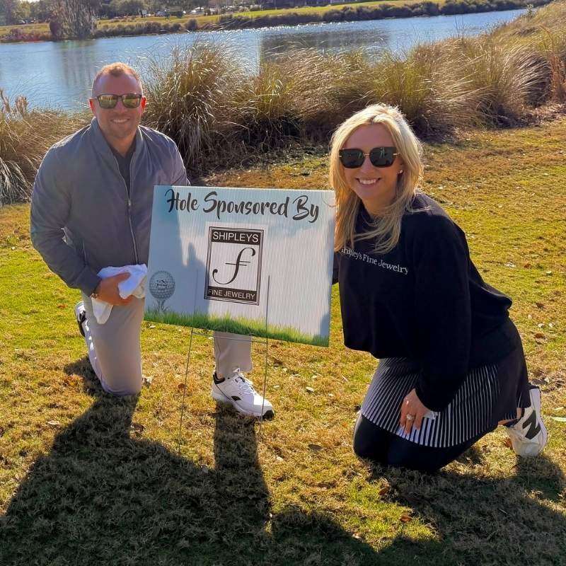 Two people kneeling on grass near a body of water, holding a sign that reads "Hole Sponsored By Shipley's Fine Jewelry," both smiling and wearing sunglasses.