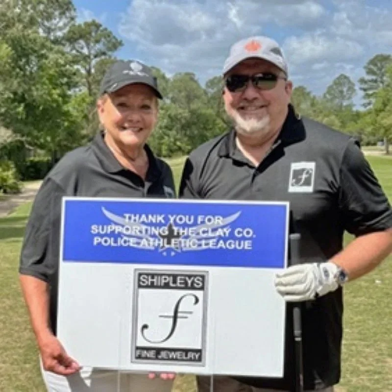 Two people in golf attire standing outdoors on a golf course, holding a sign that thanks supporters of a police athletic league, with trees and a cloudy sky in the background.